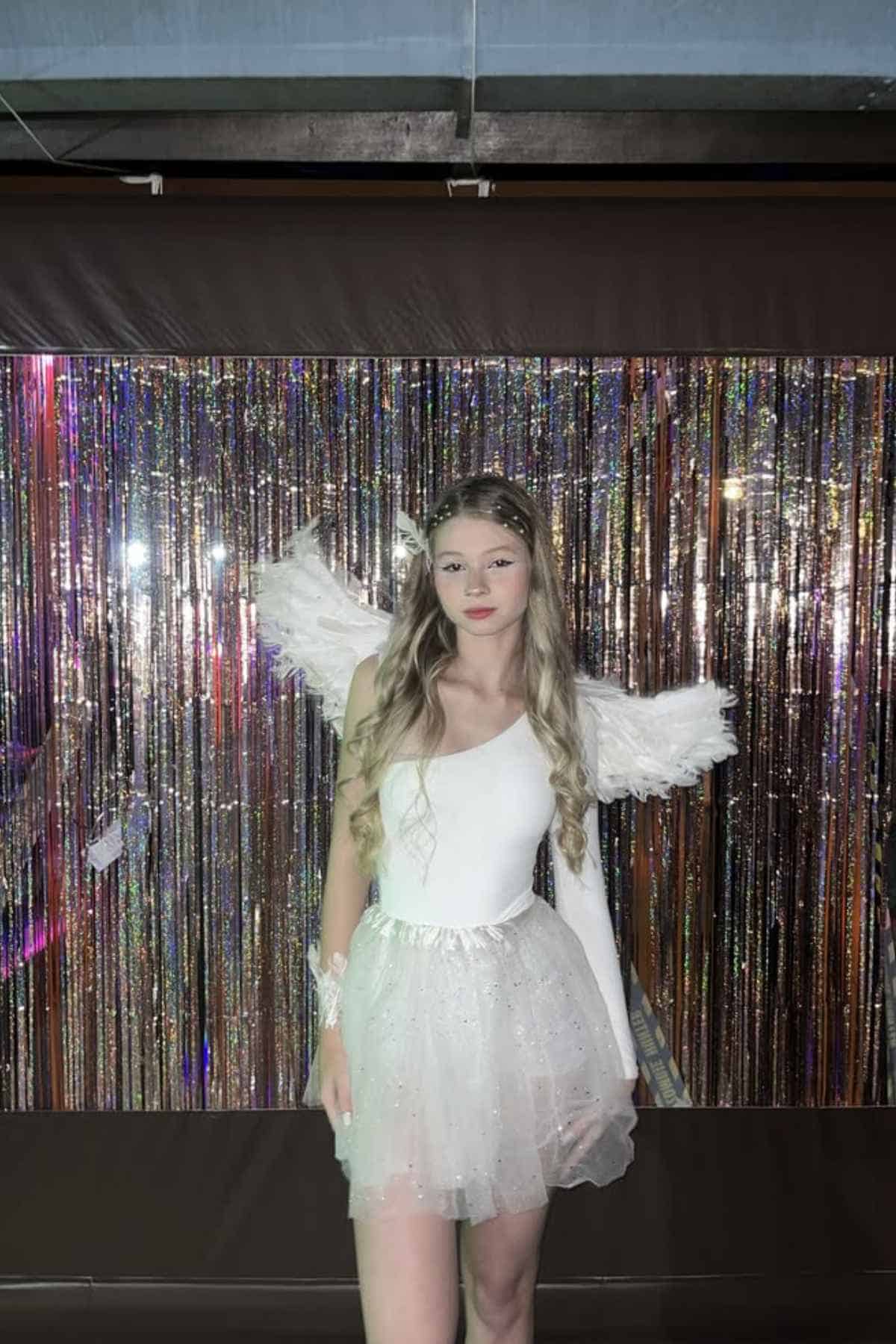 A person in a white fairy or angel costume with wings stands in front of a sparkly tinsel backdrop, looking at the camera.