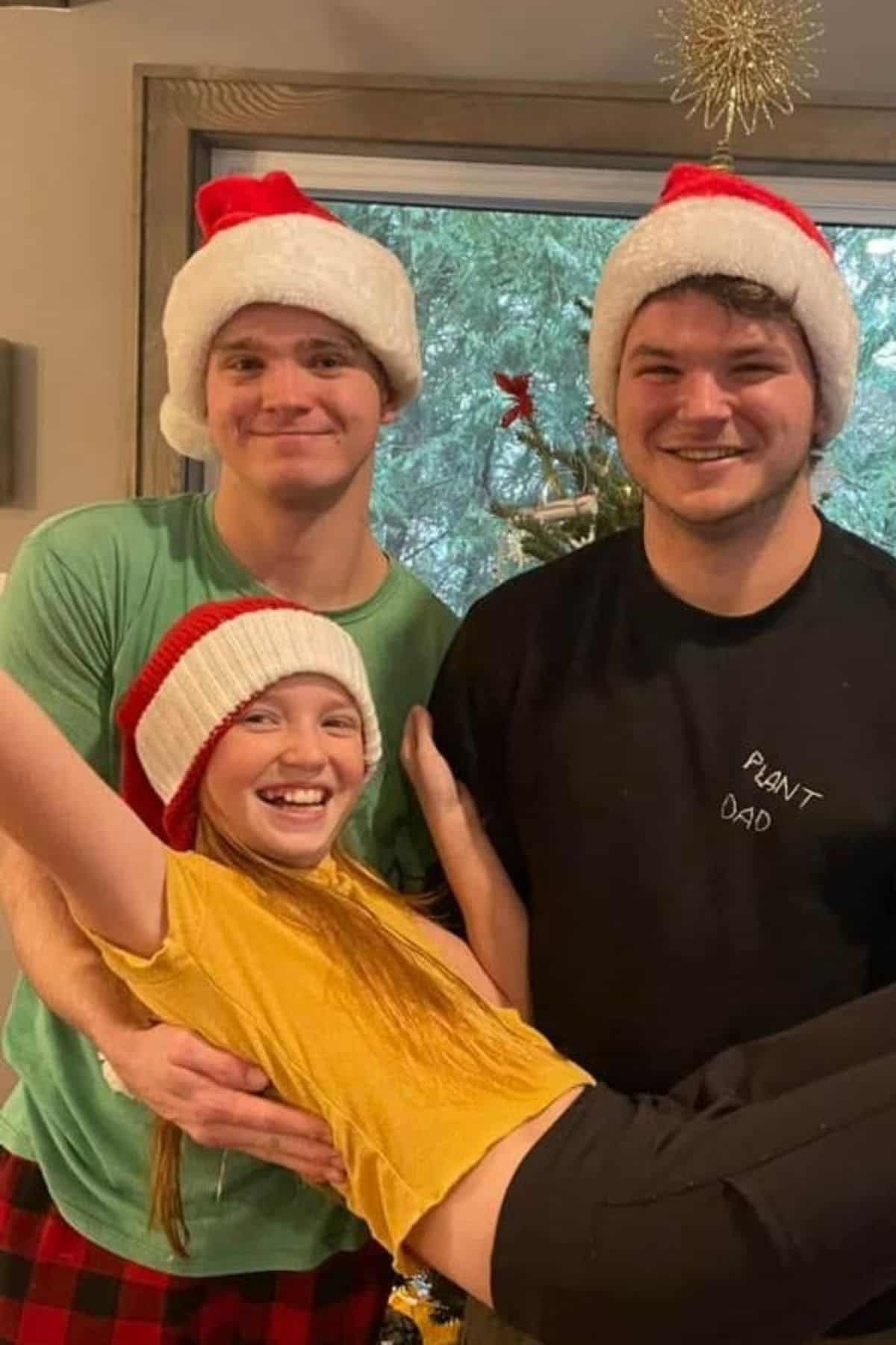Three people wearing Santa hats pose indoors; two stand while one person in front is held up horizontally, all smiling. A decorated Christmas tree is visible in the background.