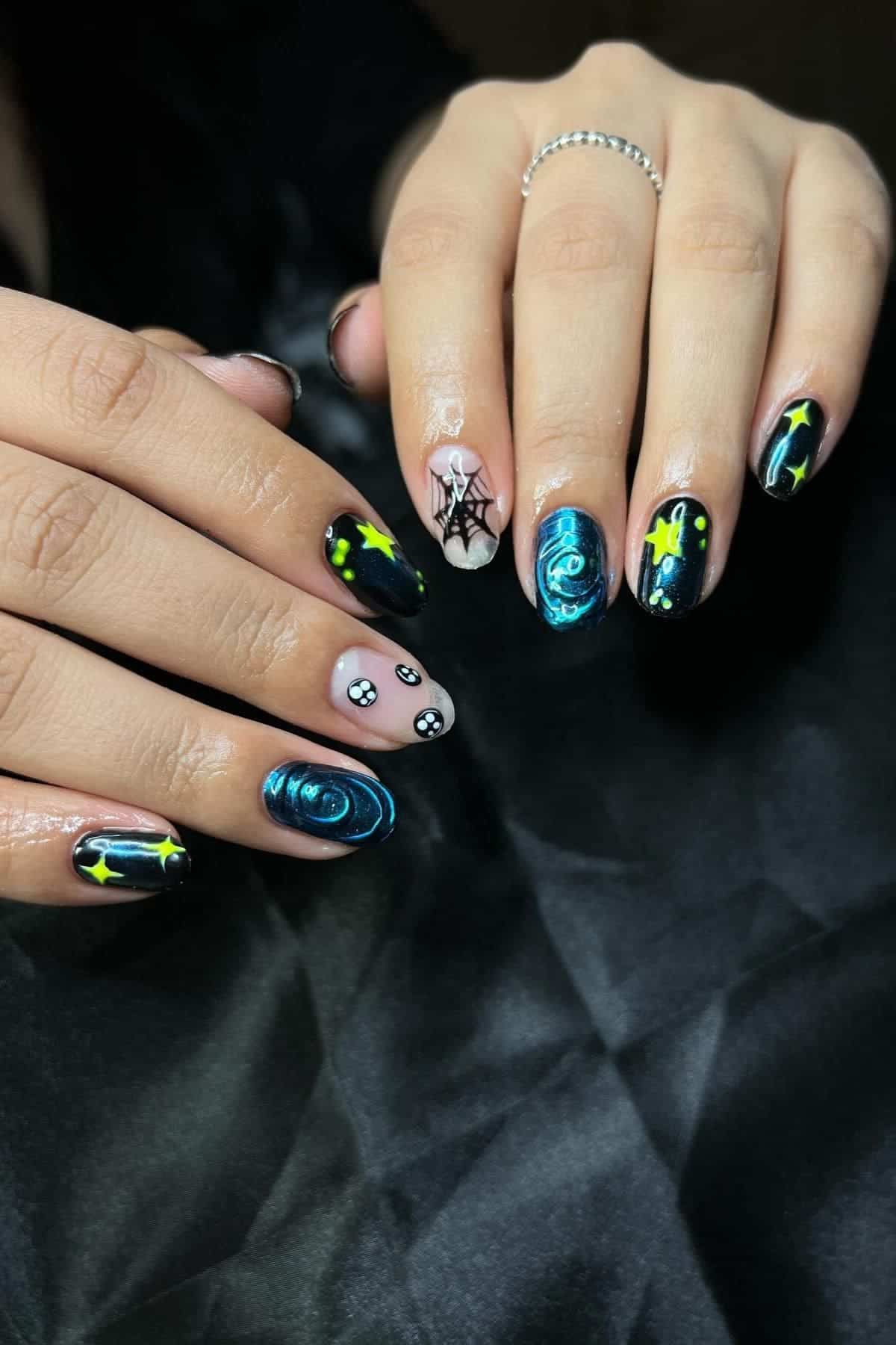 Two hands with long nails featuring a galaxy theme, including stars, planets, blue swirls, and a small spider web design on a black and nude background.