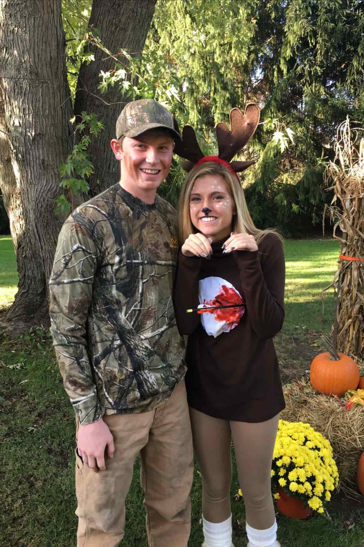 Two people pose outdoors; one wears camouflage, the other wears antlers and a brown outfit with a fake arrow and black nose, resembling a deer. Pumpkins and flowers are nearby.