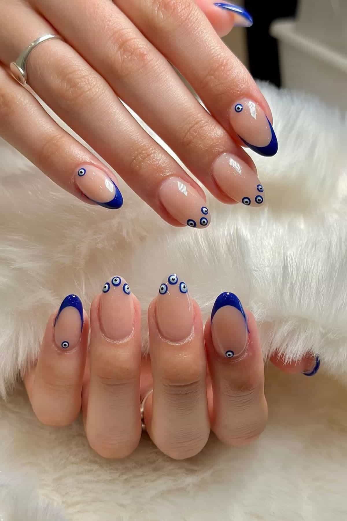Hands with almond-shaped nails featuring nude polish, blue French tips, and small blue evil eye designs on some nails, resting on a white furry surface.