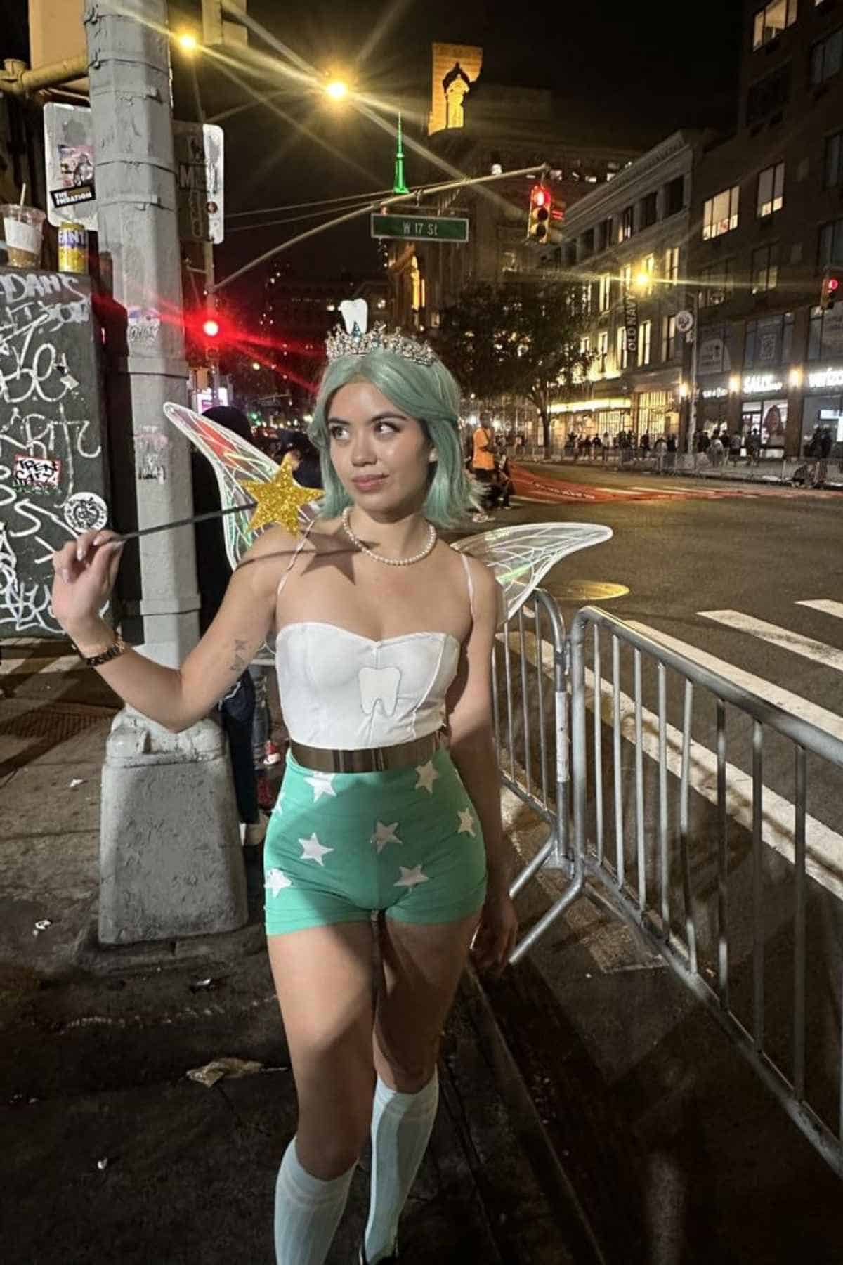 A woman in a fairy costume with wings and a crown stands on a city street at night, holding a star wand.