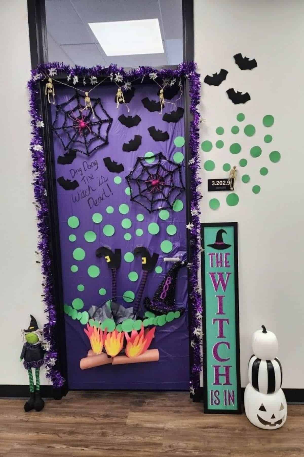 A door decorated with purple garland, bats, spider webs, witch legs, green dots, a sign reading “THE WITCH IS IN,” a pumpkin stack, and a small witch doll, Halloween-themed.