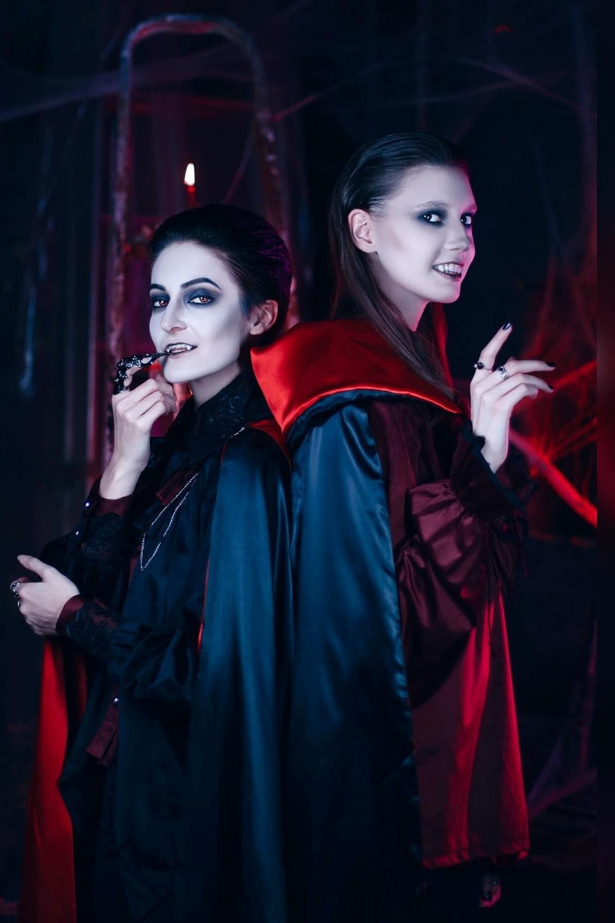 Two people in dark vampire costumes with red capes stand back to back, wearing dramatic makeup and fangs, against a dark, spooky background.