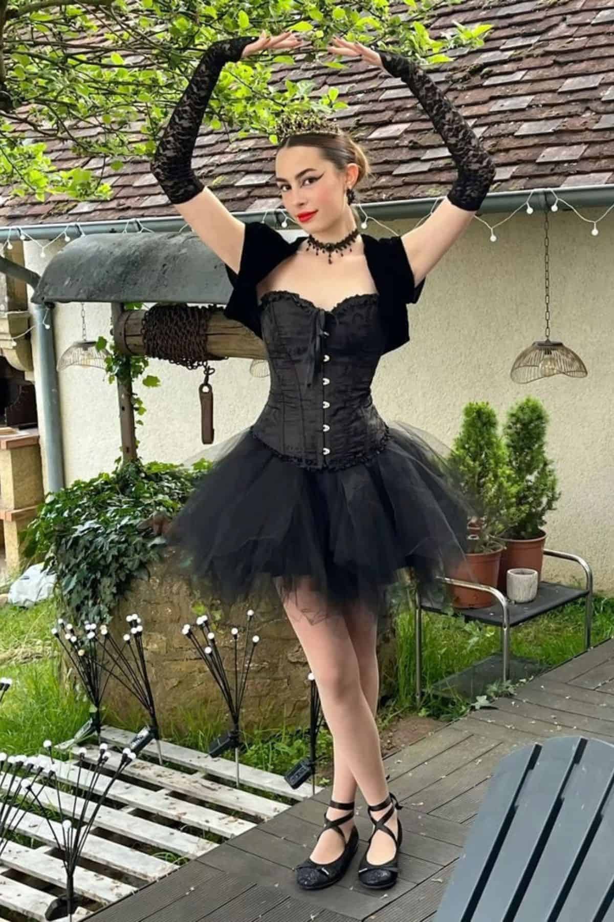 A woman in a black corset and tutu poses with arms raised in a garden, wearing lace gloves, choker necklace, and ballet-inspired shoes.
