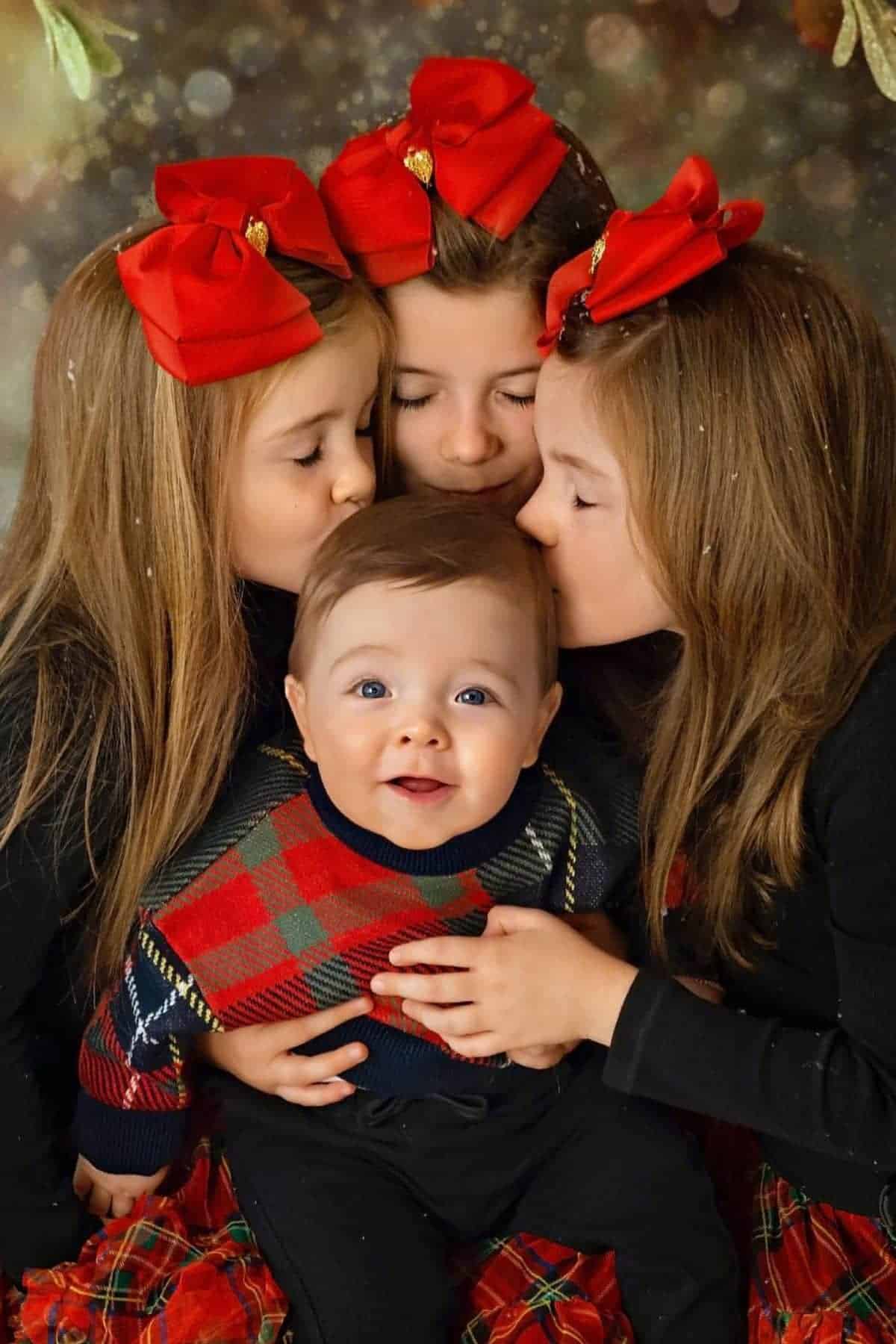 Three girls with red bows kiss the forehead of a smiling baby boy wearing a plaid sweater, posing for a holiday-themed portrait.