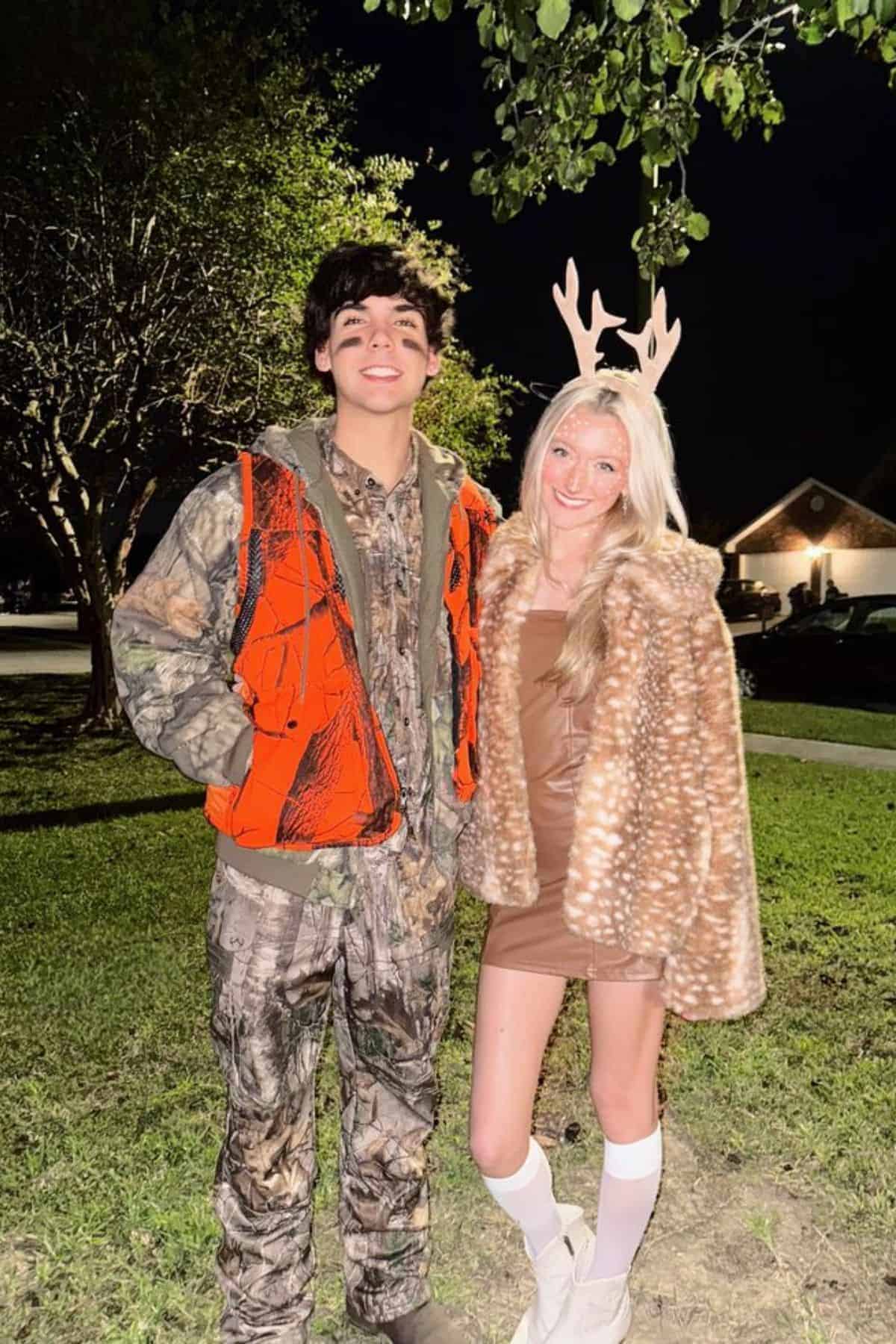 Two people pose outdoors at night; one wears camouflage with an orange vest, and the other wears a brown dress, faux fur coat, and antler headband, resembling a hunter and a deer.