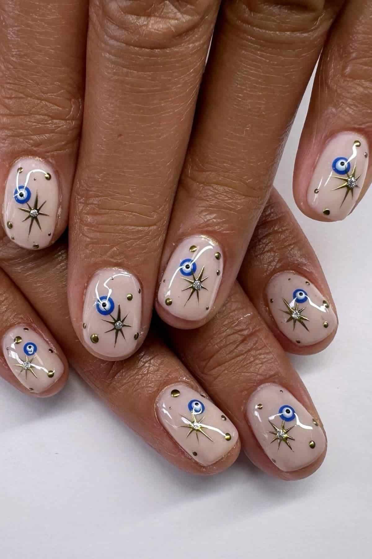 A hand with short, nude-colored nails decorated with blue evil eye designs, black stars, and small gold dots on each nail.