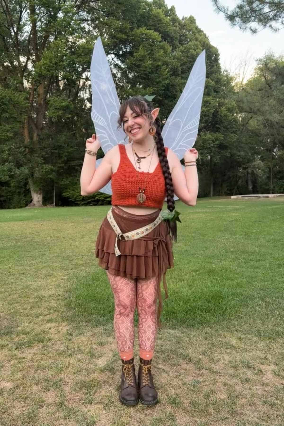 Person in a fairy costume with translucent wings, braided hair, and layered clothing stands smiling on grass in a park with trees in the background.