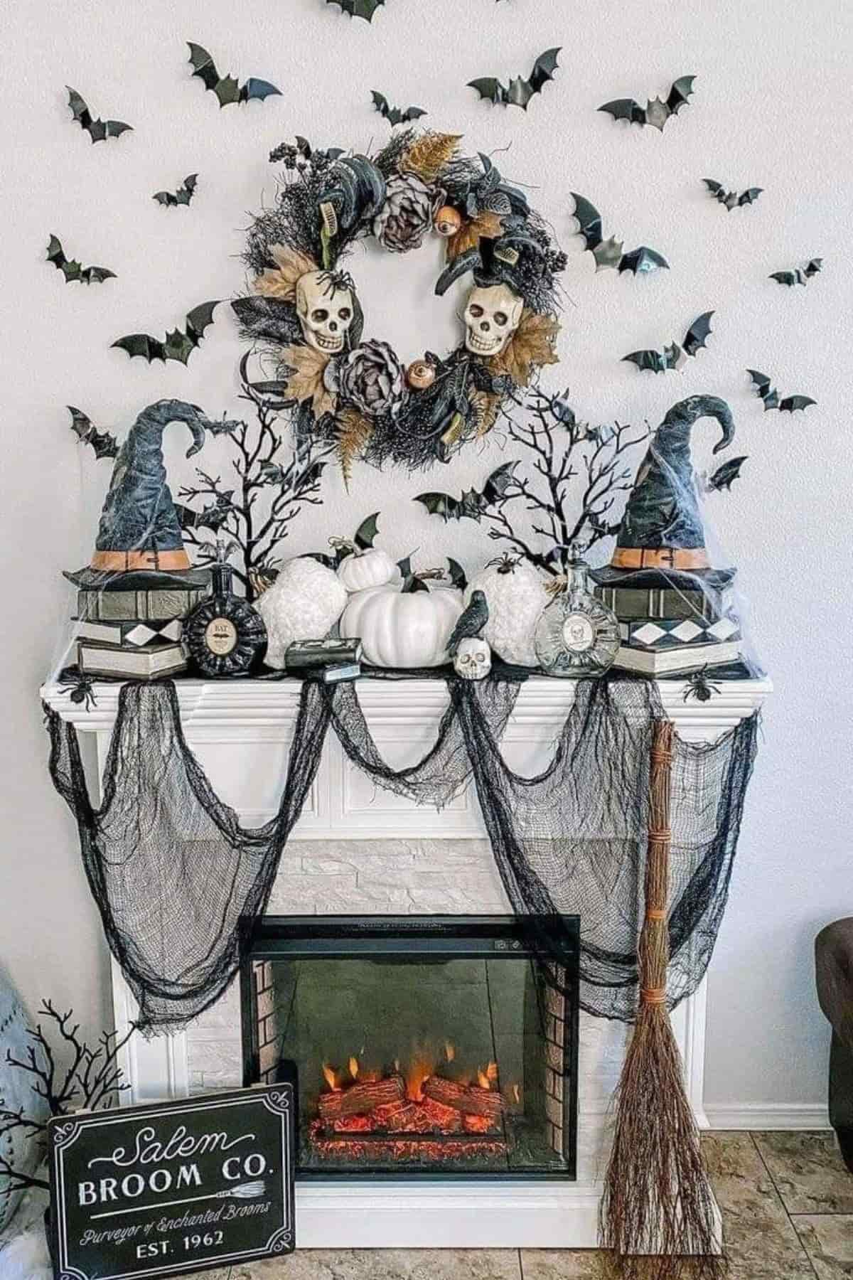 A decorated mantel features pumpkins, skulls, witch hats, faux black trees, a broom, bats on the wall, and a wreath with skulls above a lit electric fireplace.