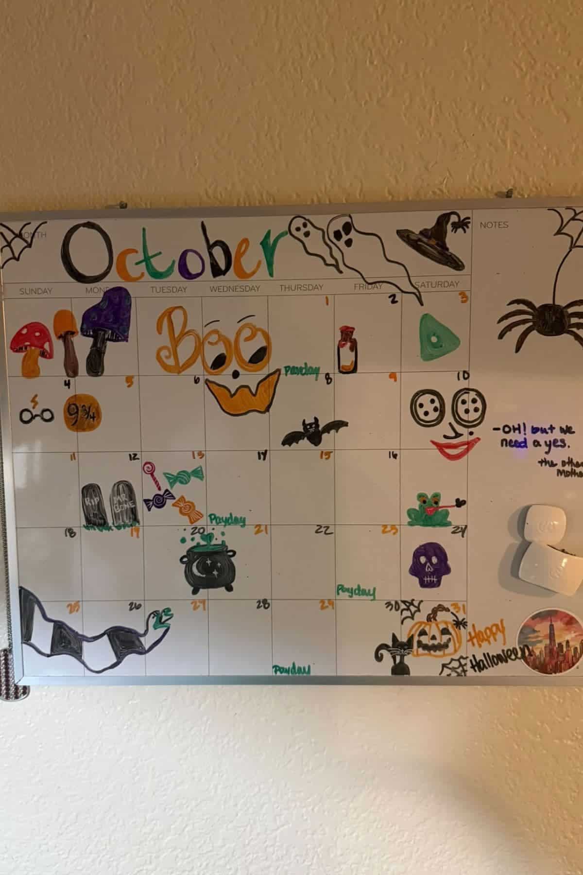 A whiteboard calendar for October is decorated with hand-drawn Halloween-themed images including ghosts, pumpkins, and spiders. Some days are labeled “Payday” and “Halloween.”.