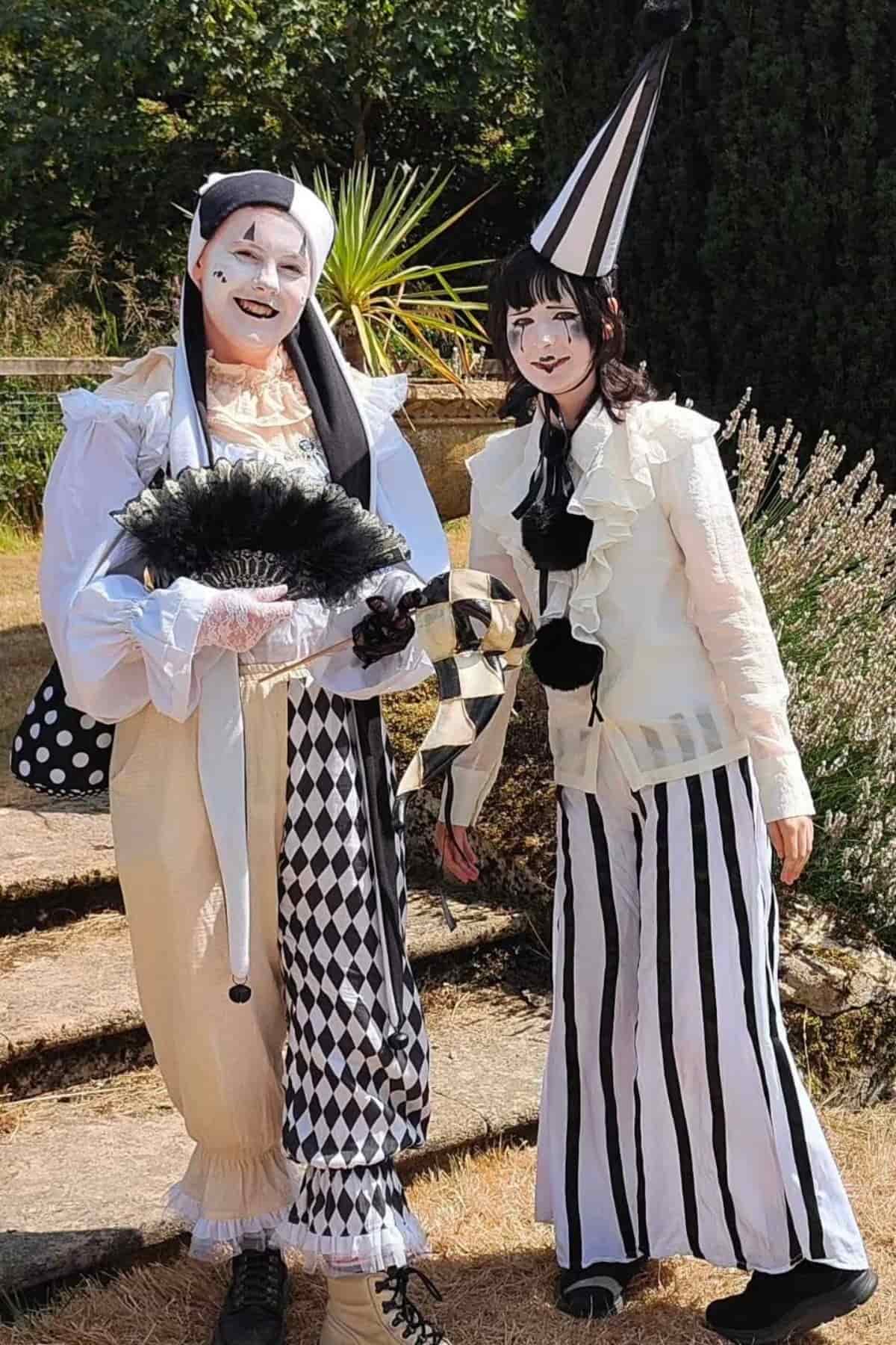 Two people in black and white harlequin costumes stand outdoors, one holding a fan and the other with a tall pointed hat; both have painted faces and ruffled collars.
