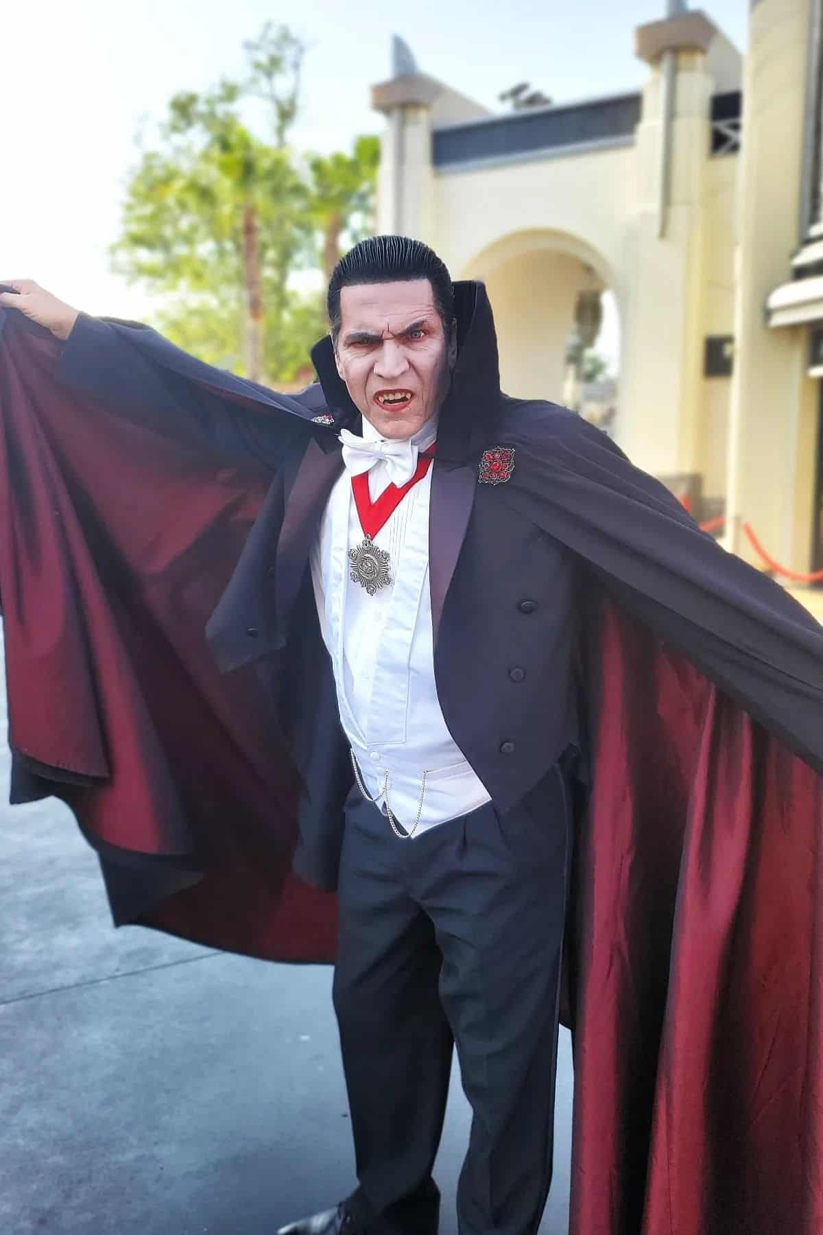 Person dressed as a classic vampire in a black cape with red lining, white shirt, and medallion, standing outdoors with arms spread wide.