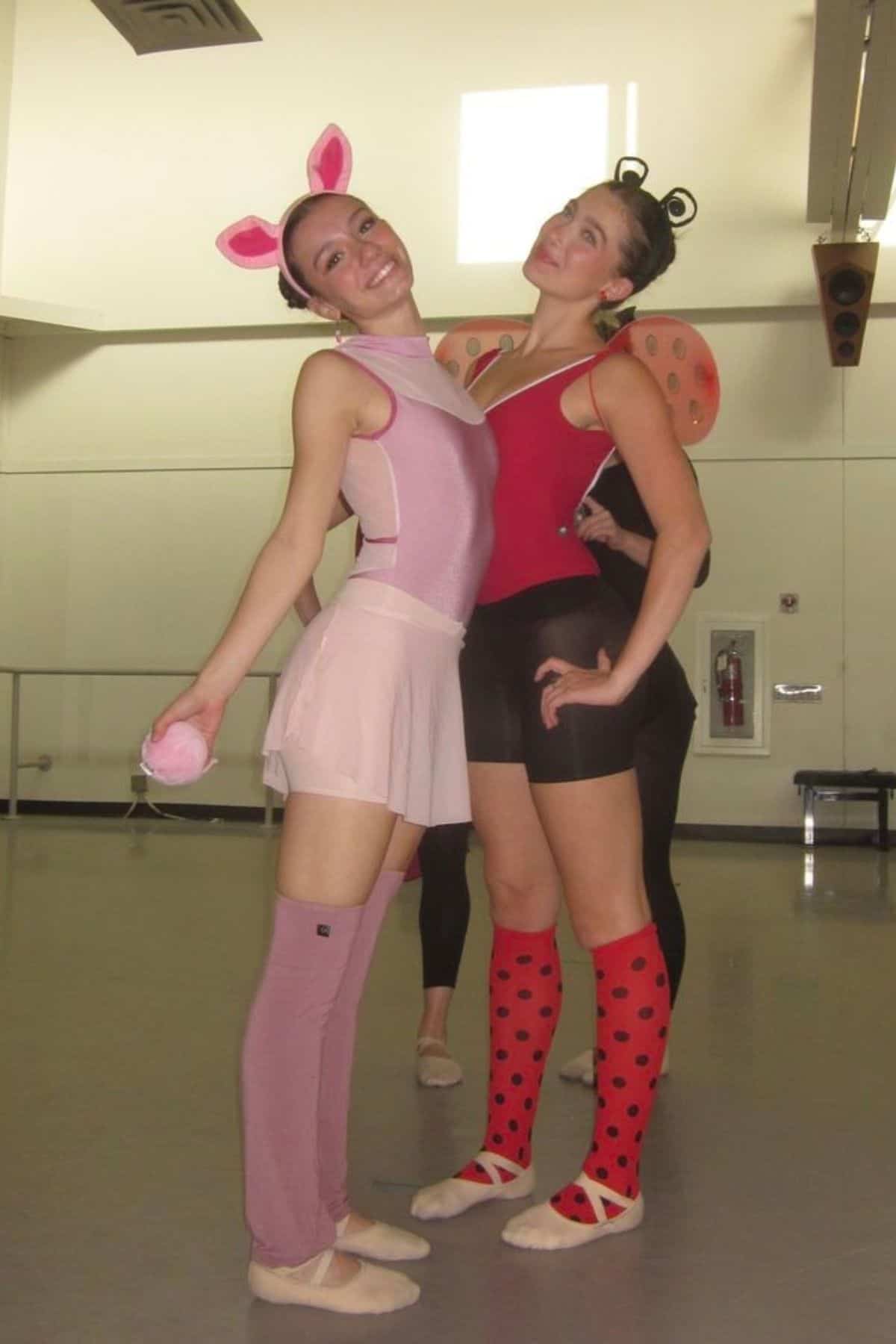 Two ballet dancers pose in costume; one wears pink with pig ears, the other wears red with ladybug wings and socks. They stand in a dance studio with mirrors and ballet barres.