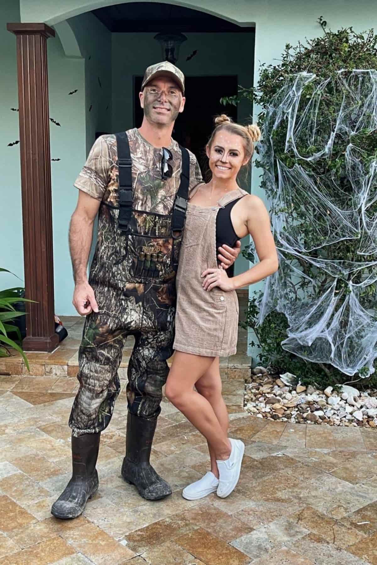 Two people pose outside; one wears camouflage overalls, boots, and a cap, while the other wears a tan overall dress and white sneakers. There are fake cobwebs on a bush nearby.
