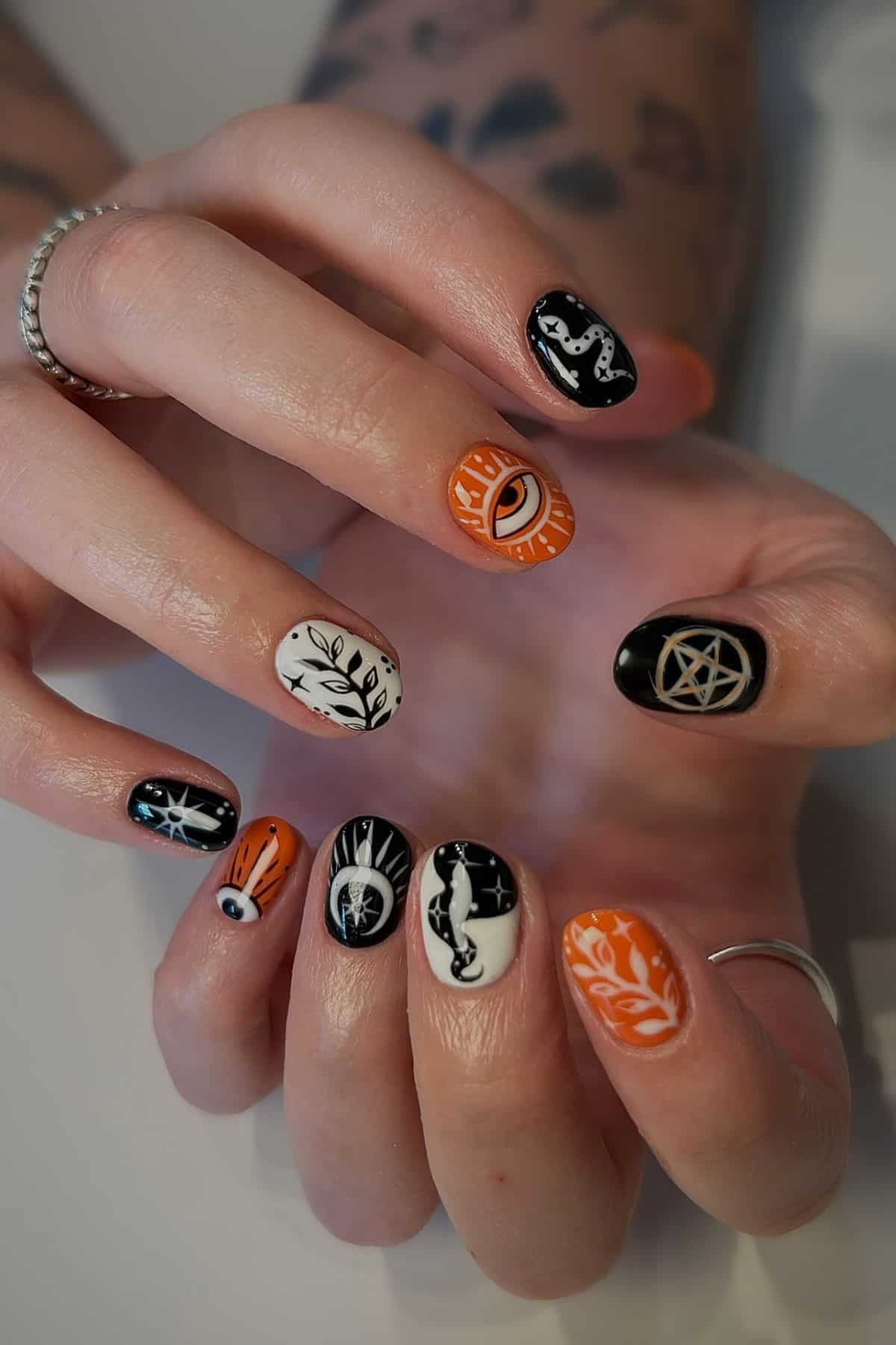 A hand with black, white, and orange nail art featuring symbols like an eye, pentagram, moon, leaves, and abstract designs.