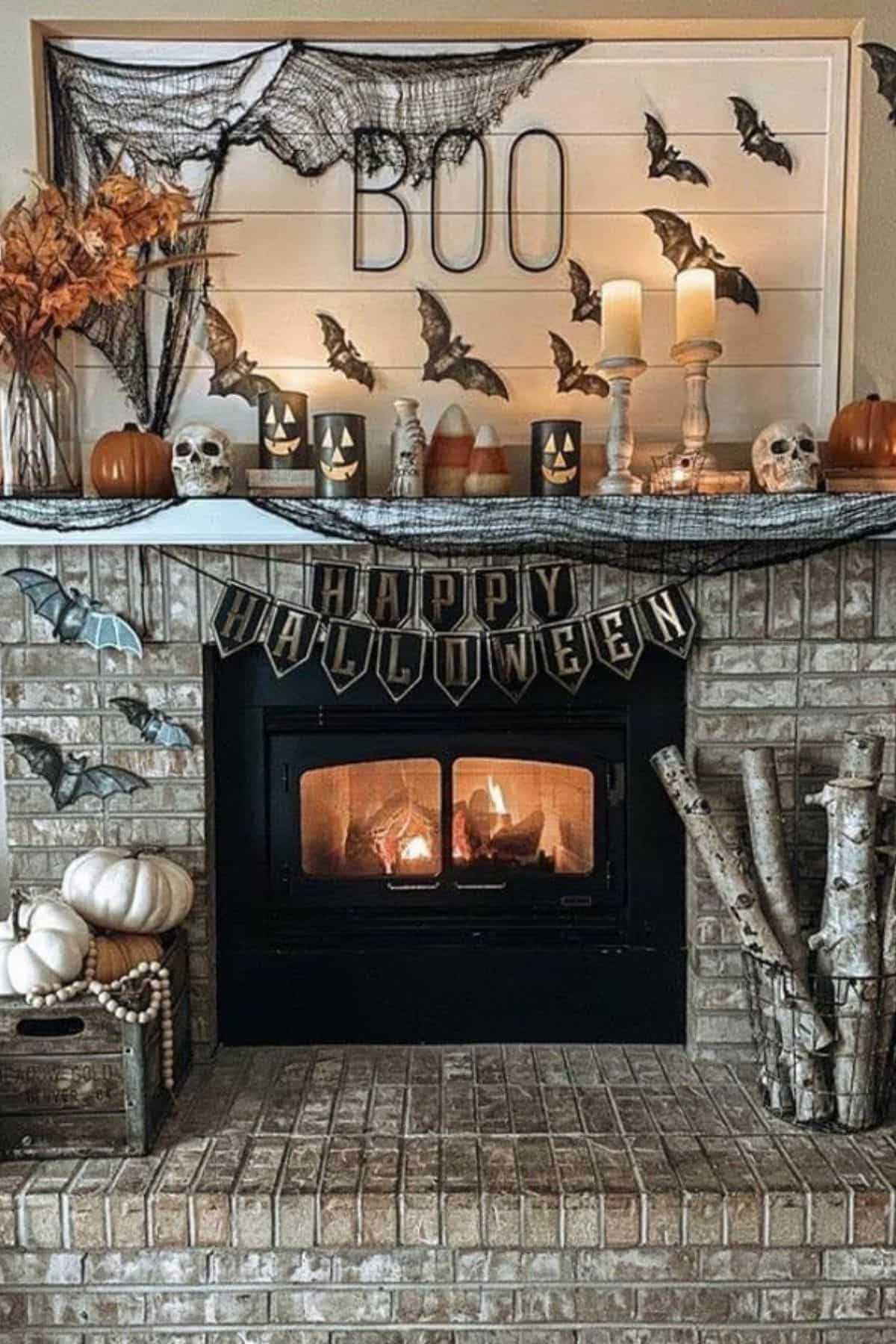 Brick fireplace decorated for Halloween with “BOO” sign, bats, skulls, candles, pumpkins, fall leaves, and “Happy Halloween” banner. Fire burns in the hearth below.