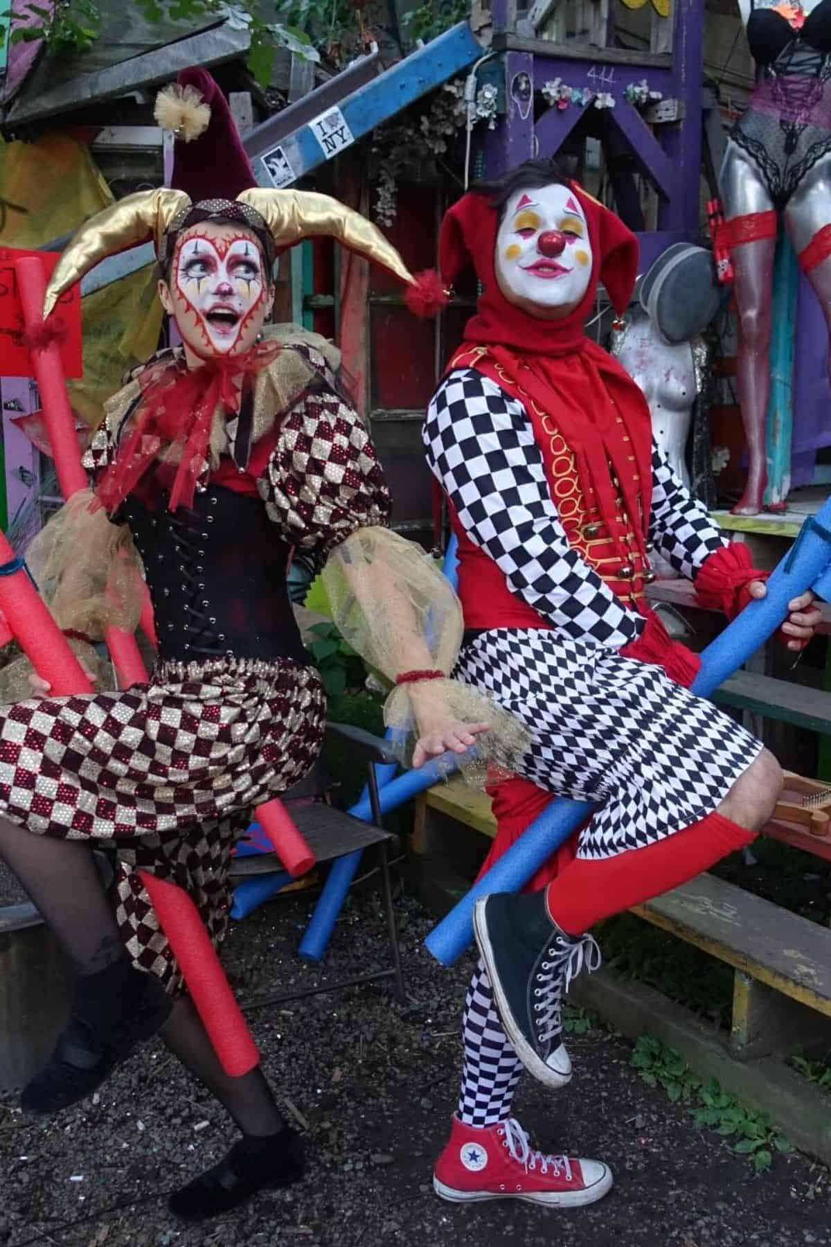 Two people in harlequin costumes pose outdoors, each with painted faces, colorful hats, and bright patterned clothing, holding blue foam sticks, surrounded by eclectic decorations and mannequins.