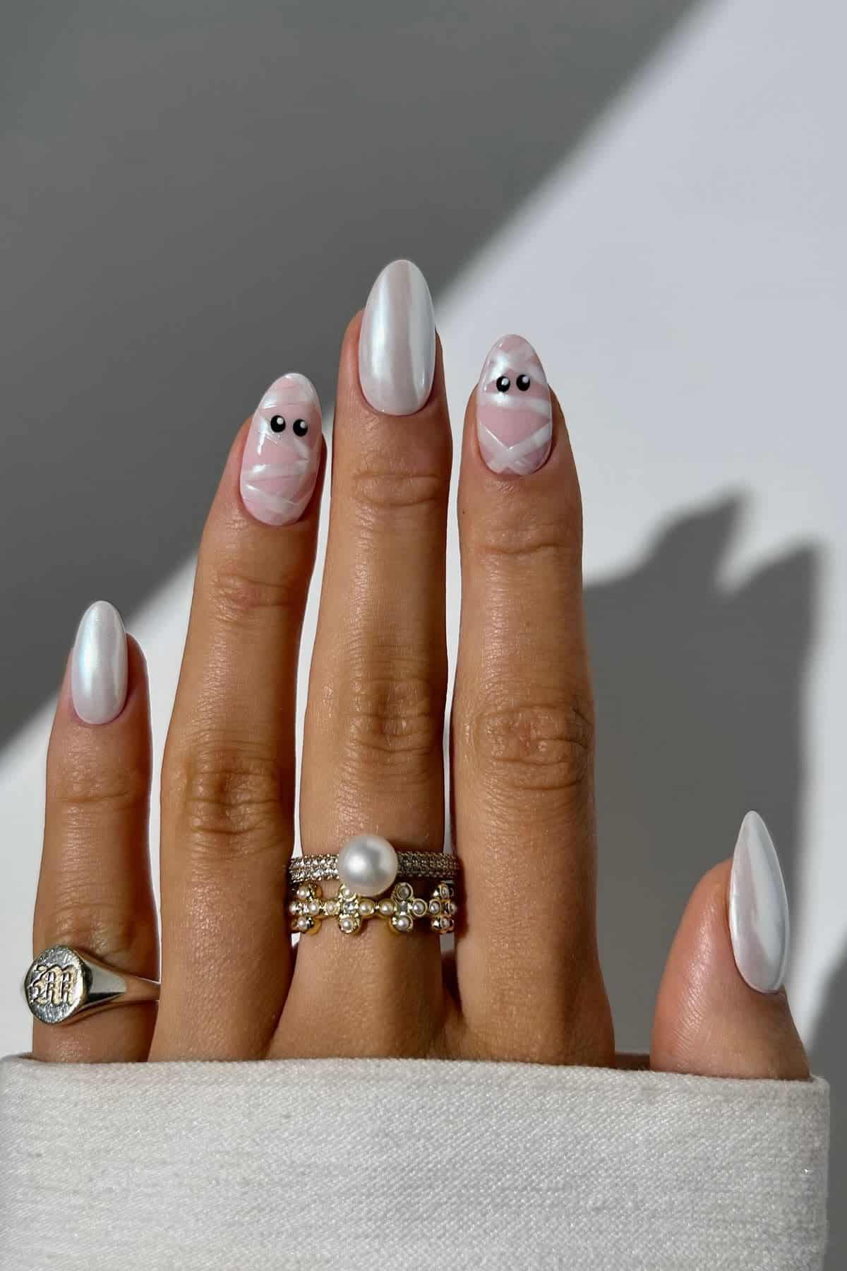 A hand with long, pearl-white nails featuring two accent nails with mummy designs and black dot eyes, wearing silver rings.