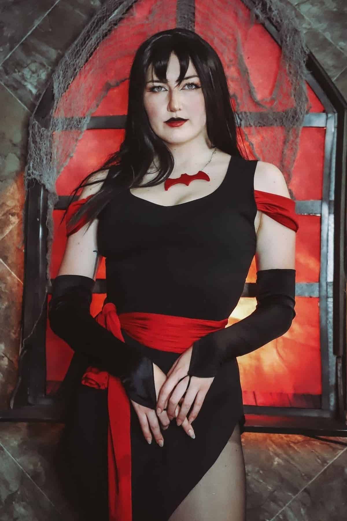 Person in a black and red costume stands in front of a gothic window with red lighting and cobweb decorations.