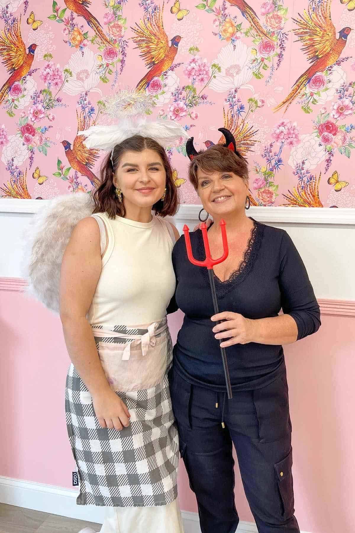 Two women stand smiling; one is dressed as an angel with wings and a halo, the other as a devil with red horns and a pitchfork, against a floral pink wallpaper background.
