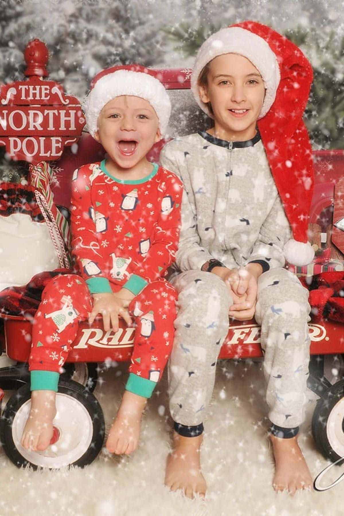 Two children in festive pajamas and Santa hats sit on a red wagon labeled “NORTH POLE,” surrounded by falling snow in a holiday-themed setting.
