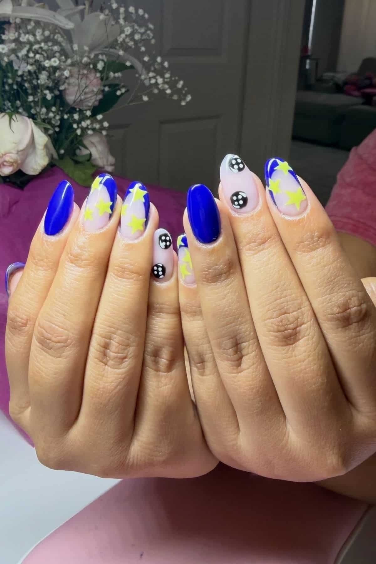 Hands with long nails painted blue and nude, decorated with yellow stars and black-and-white smiley faces; flowers visible in the background.