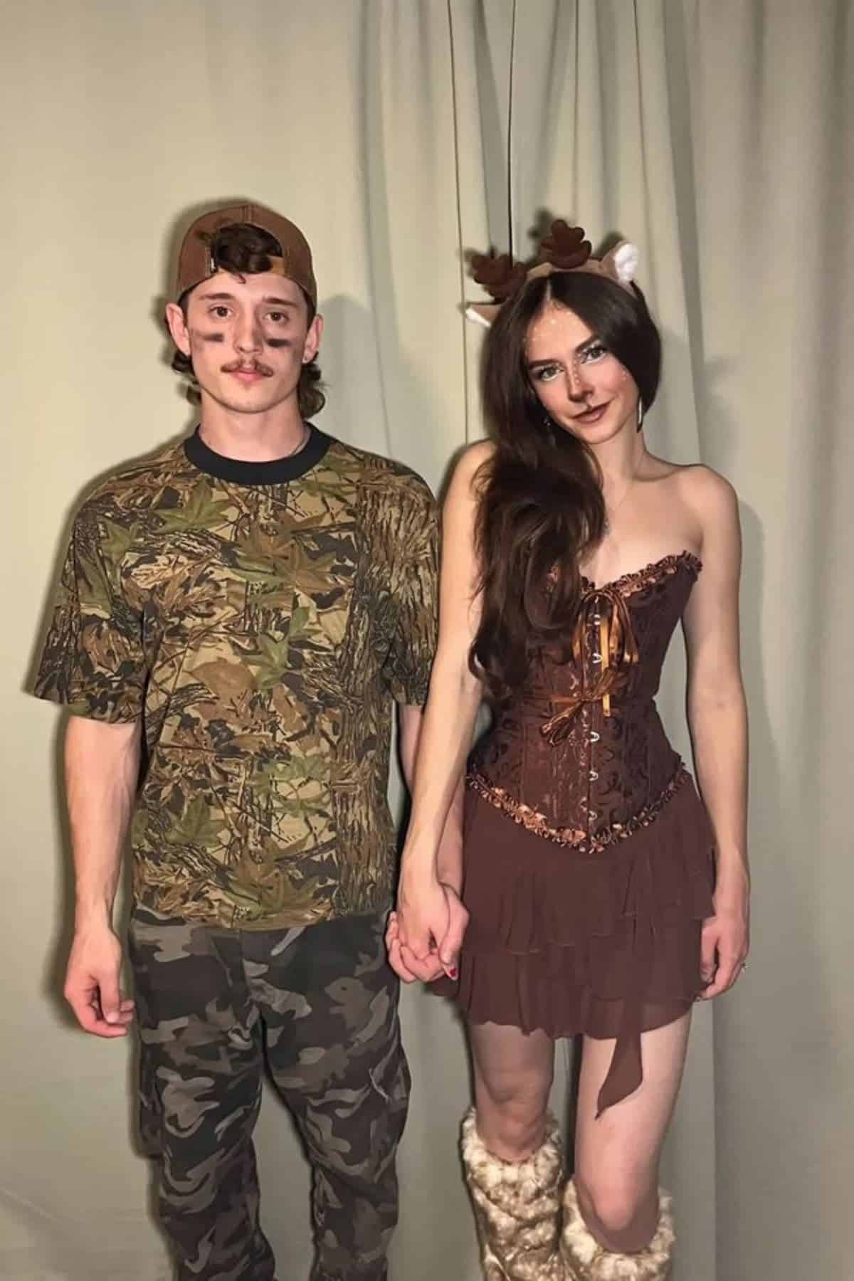 Two people stand holding hands, one dressed in camouflage hunter attire and the other in a brown costume with antlers and faux fur boots, posing in front of a plain curtain background.