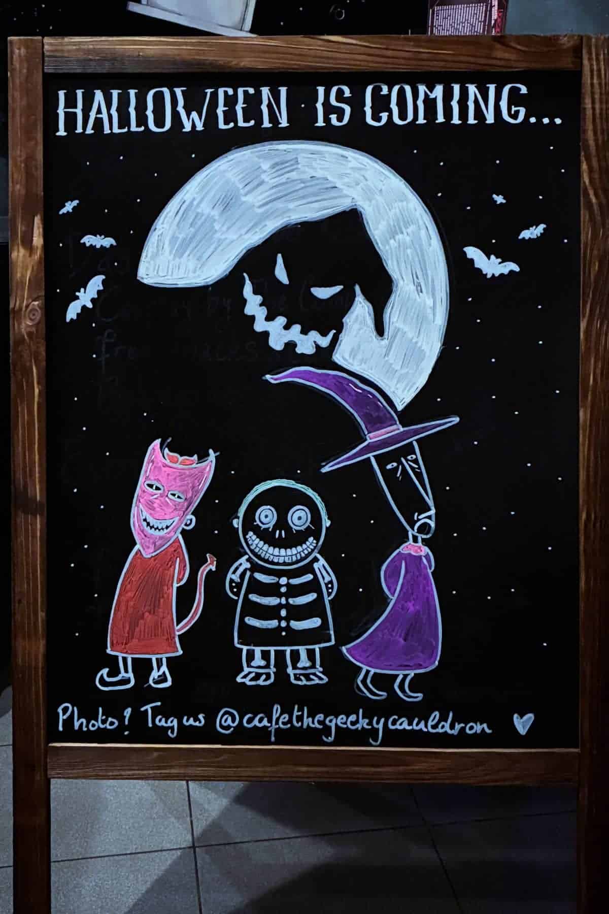Chalkboard sign with “Halloween is coming,” a spooky moon, bats, and three cartoon characters in costume: a devil, skeleton, and witch.