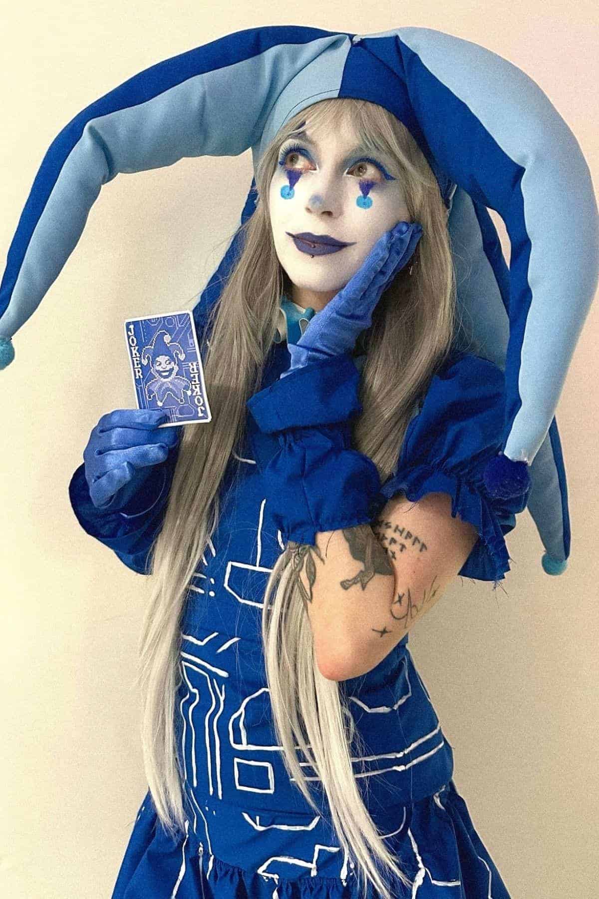 Person in a blue and light blue jester costume with matching makeup holds a blue card while posing against a plain background.