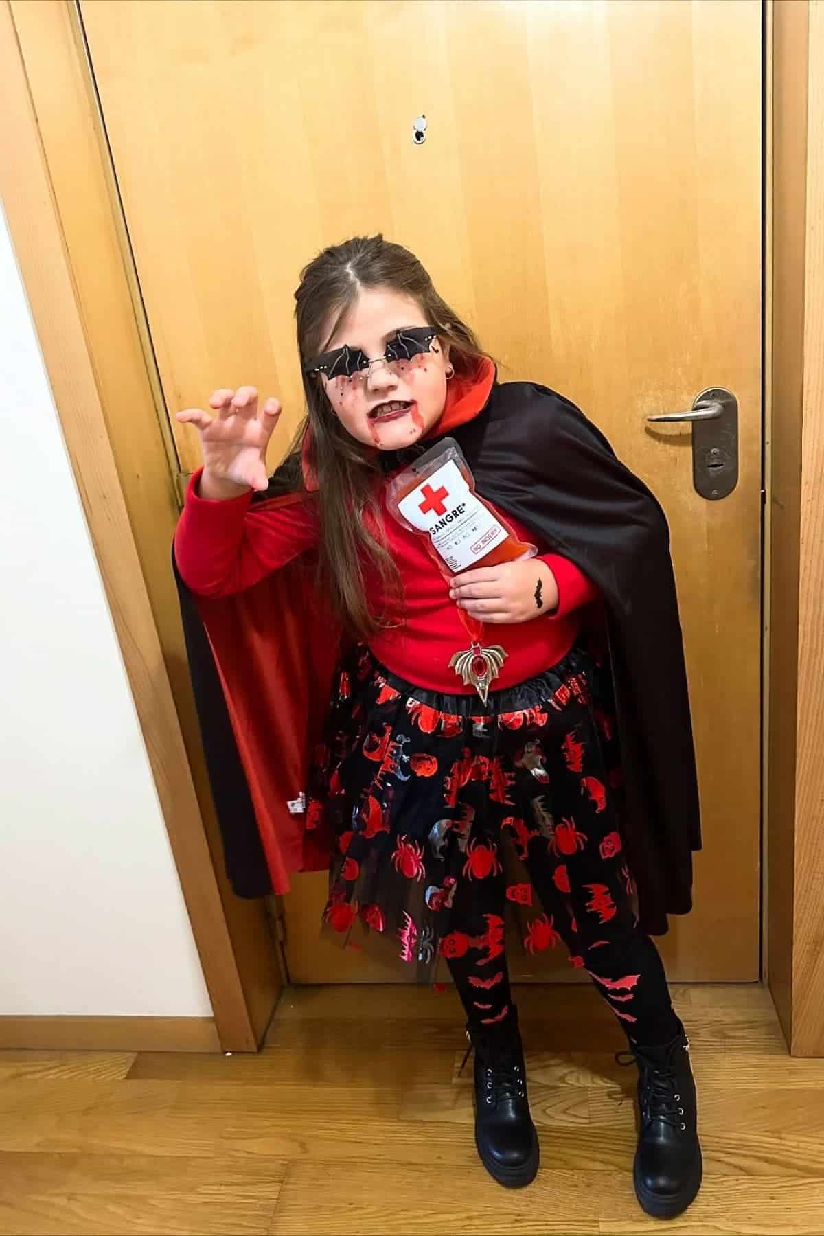 Child in a vampire costume poses indoors, wearing a black cape, red shirt, themed skirt, face paint, and holding a fake blood bag.