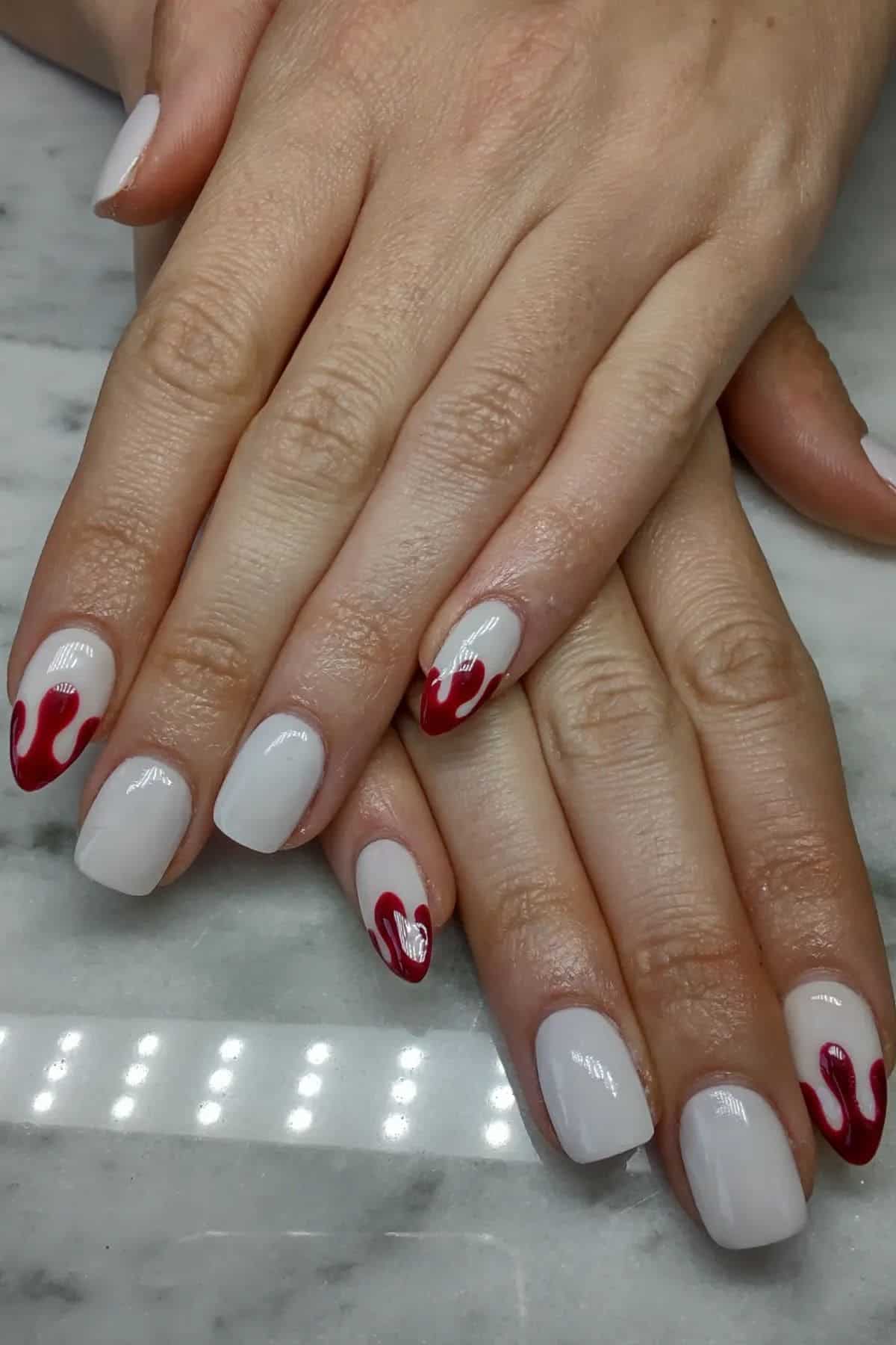 Hands with short, square-shaped white nails. Accent nails feature red flame designs on ring fingers. Nails rest on a marble surface.