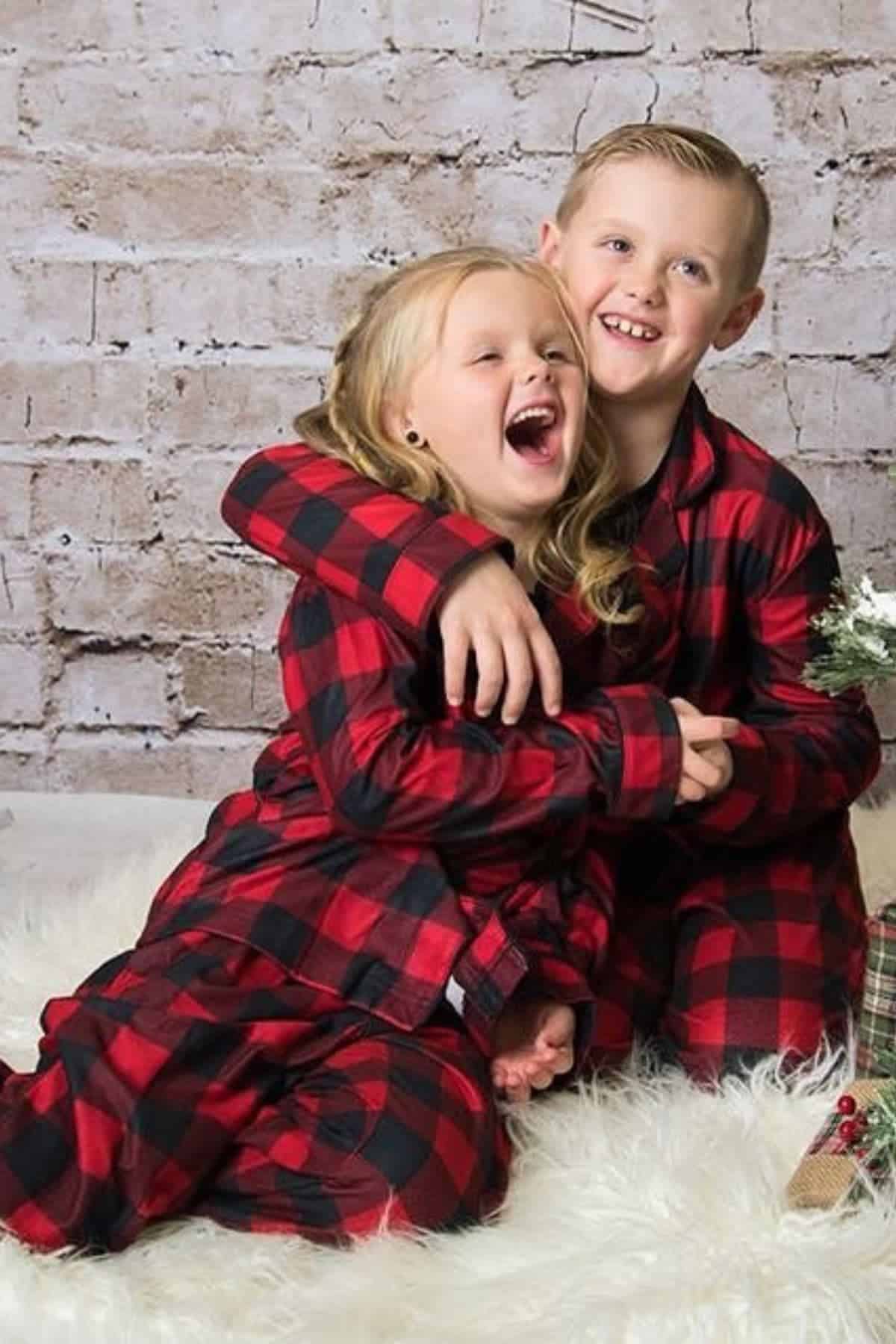 Two children wearing matching red and black plaid pajamas sit on a white rug, smiling and hugging in front of a brick wall backdrop.