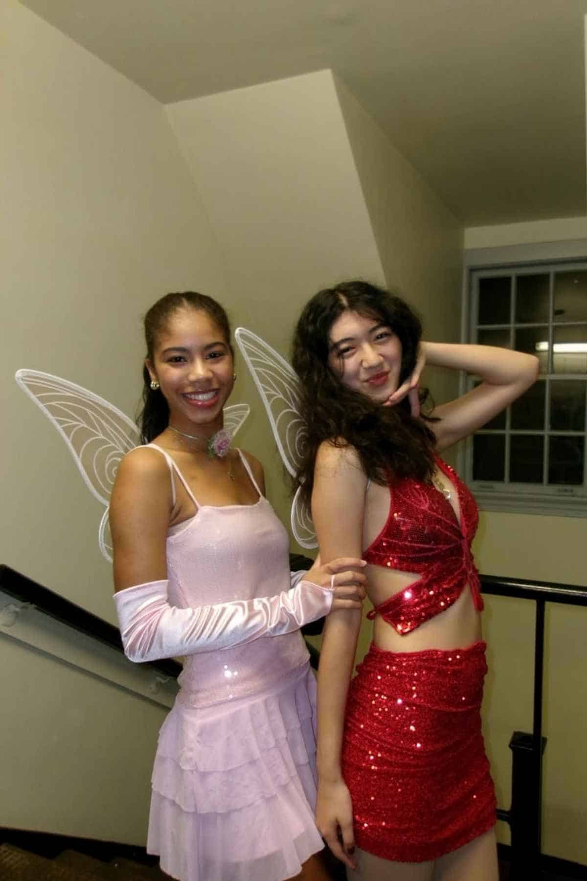 Two young women pose indoors; one wears a pink fairy costume with wings and gloves, the other wears a red sequin outfit with a cutout midriff.
