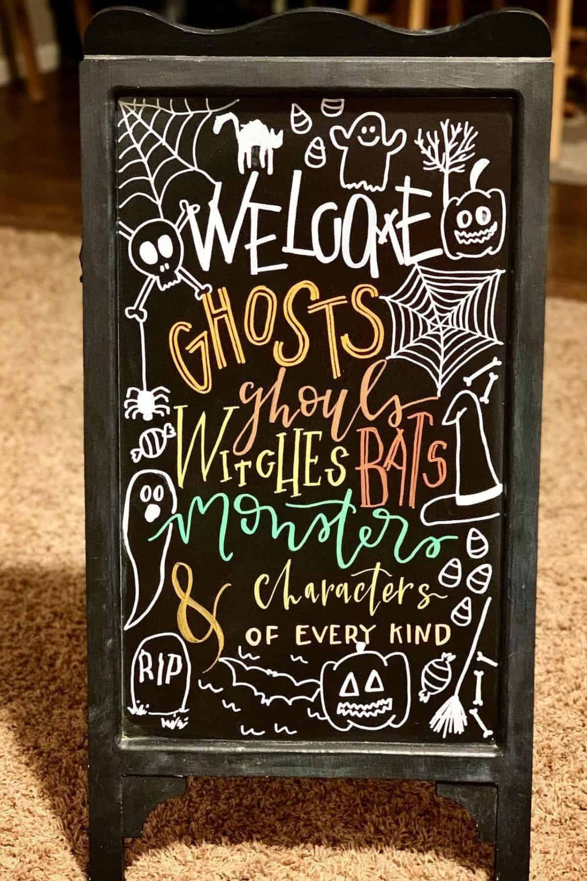 Chalkboard sign with Halloween drawings and text welcoming ghosts, ghouls, witches, bats, monsters, and characters of every kind.