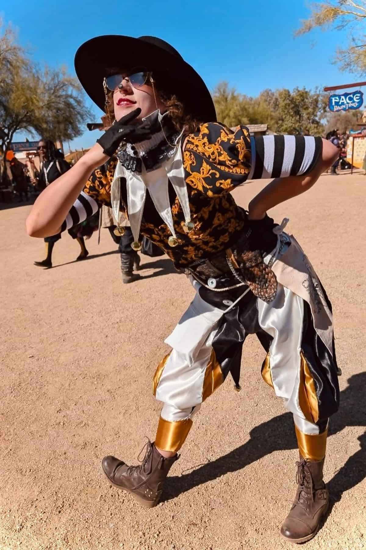 Person in a decorative costume with striped sleeves, gold accents, boots, and a wide-brimmed hat poses outdoors on a sunny day.