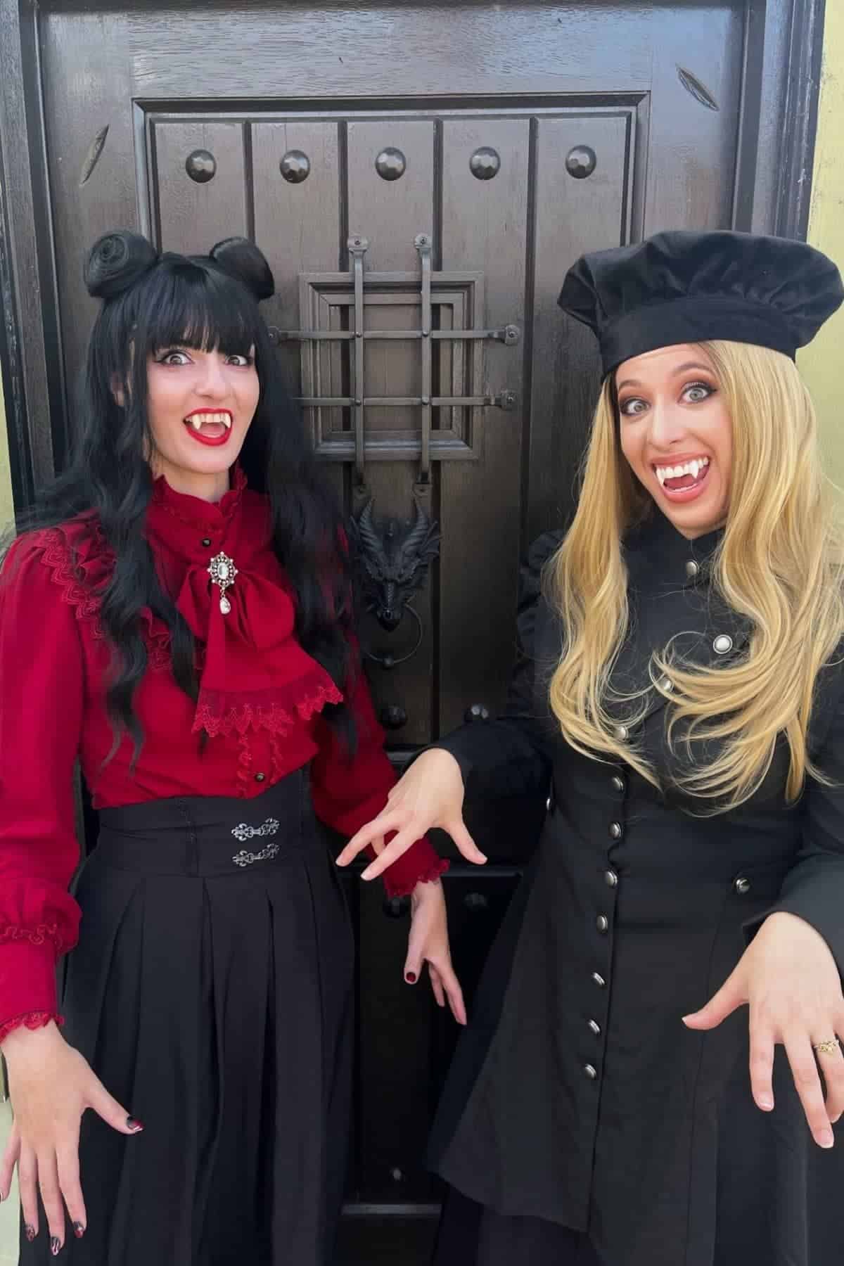 Two women in vintage-style clothing pose playfully with vampire fangs in front of a dark wooden door.