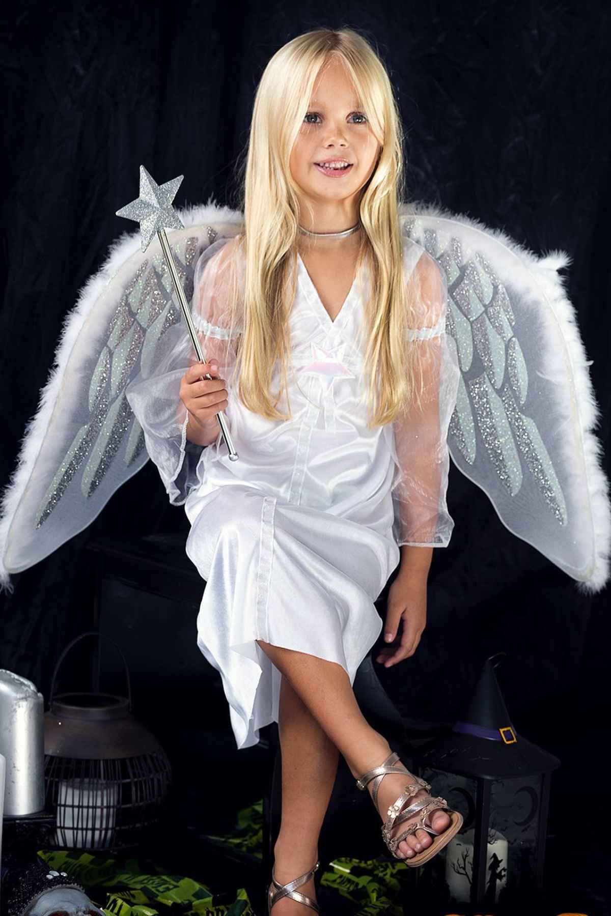 A young girl wearing angel wings and a white dress holds a silver star wand, sitting in front of a dark background with decorative items around her.