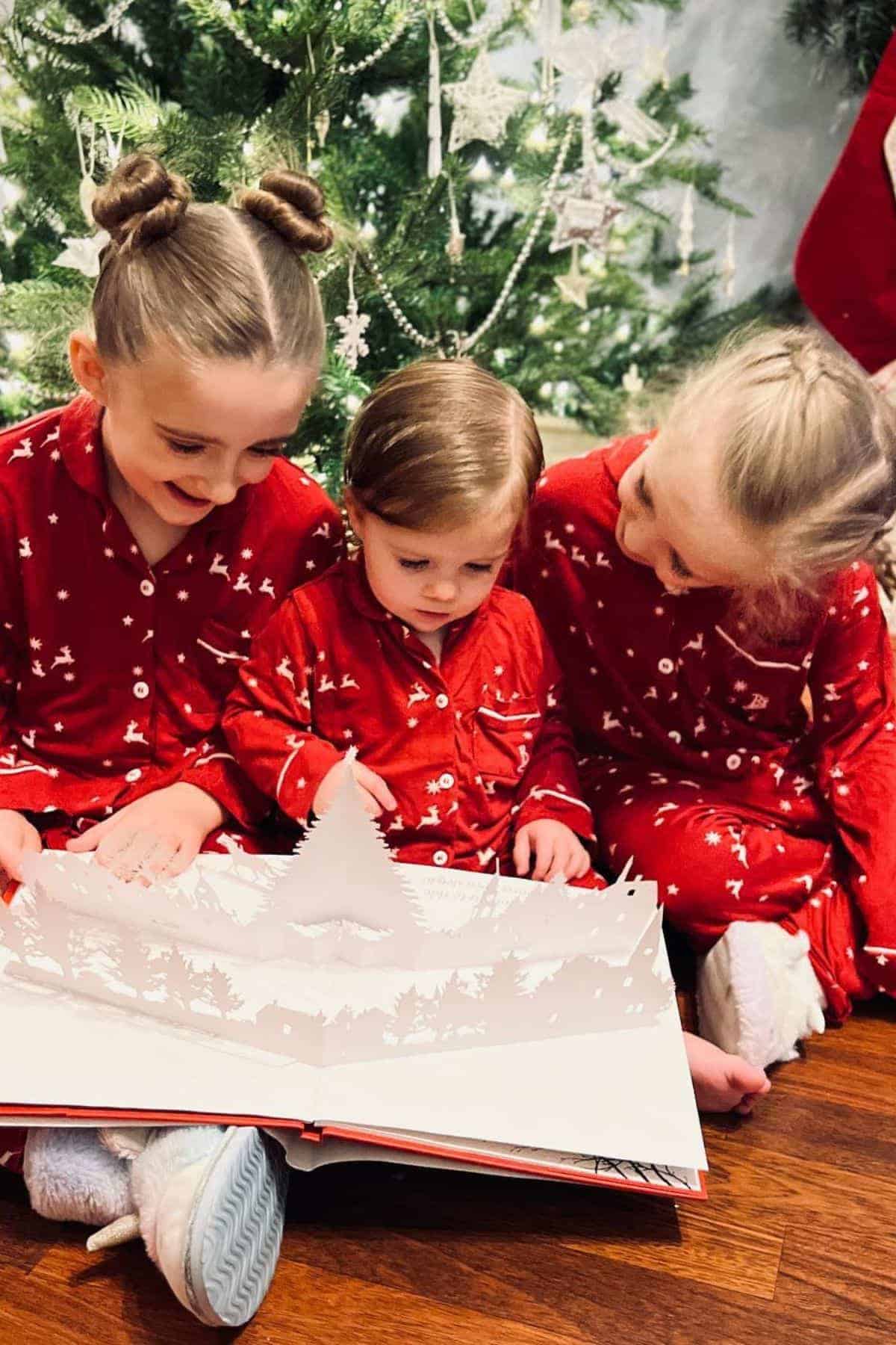 Three children in matching red pajamas sit on the floor in front of a Christmas tree, looking at a pop-up book together.