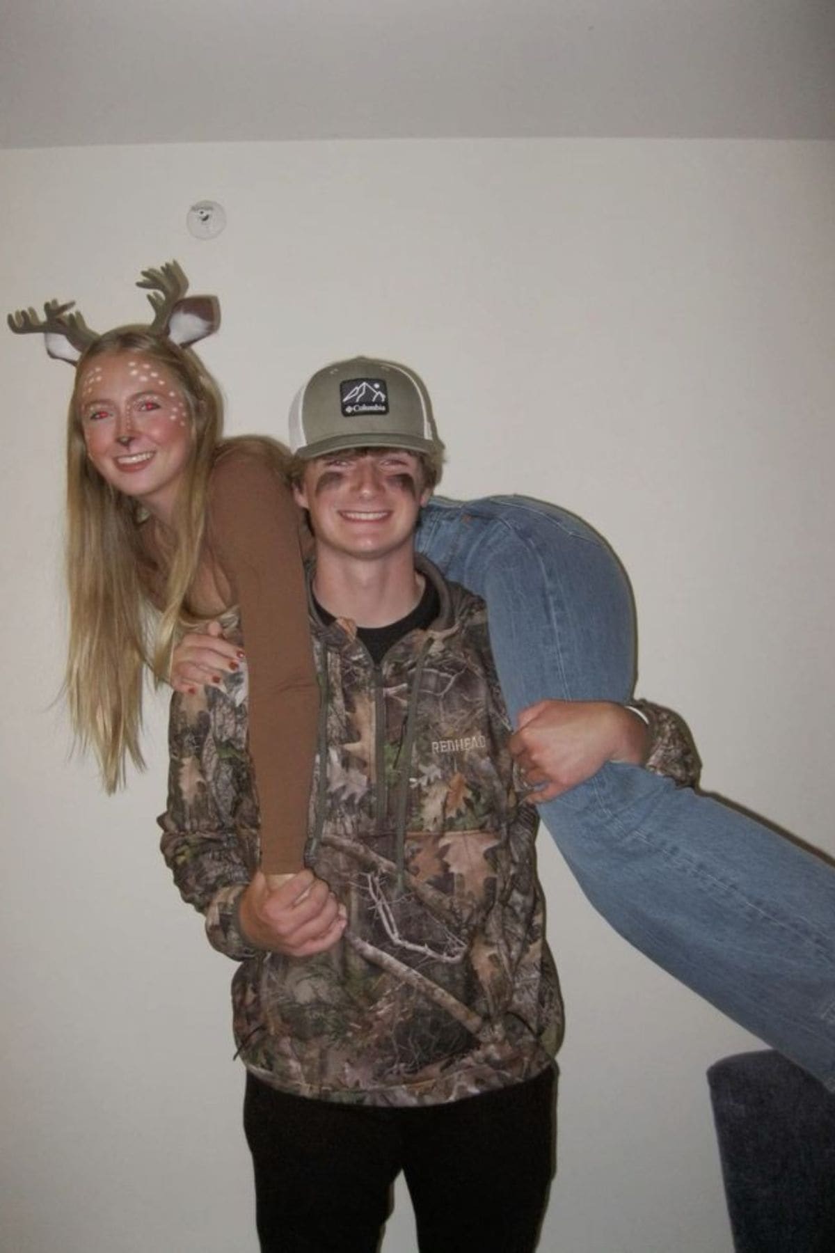A young woman in deer makeup and antlers is carried over the shoulder of a smiling young man in camouflage clothing and a baseball cap.