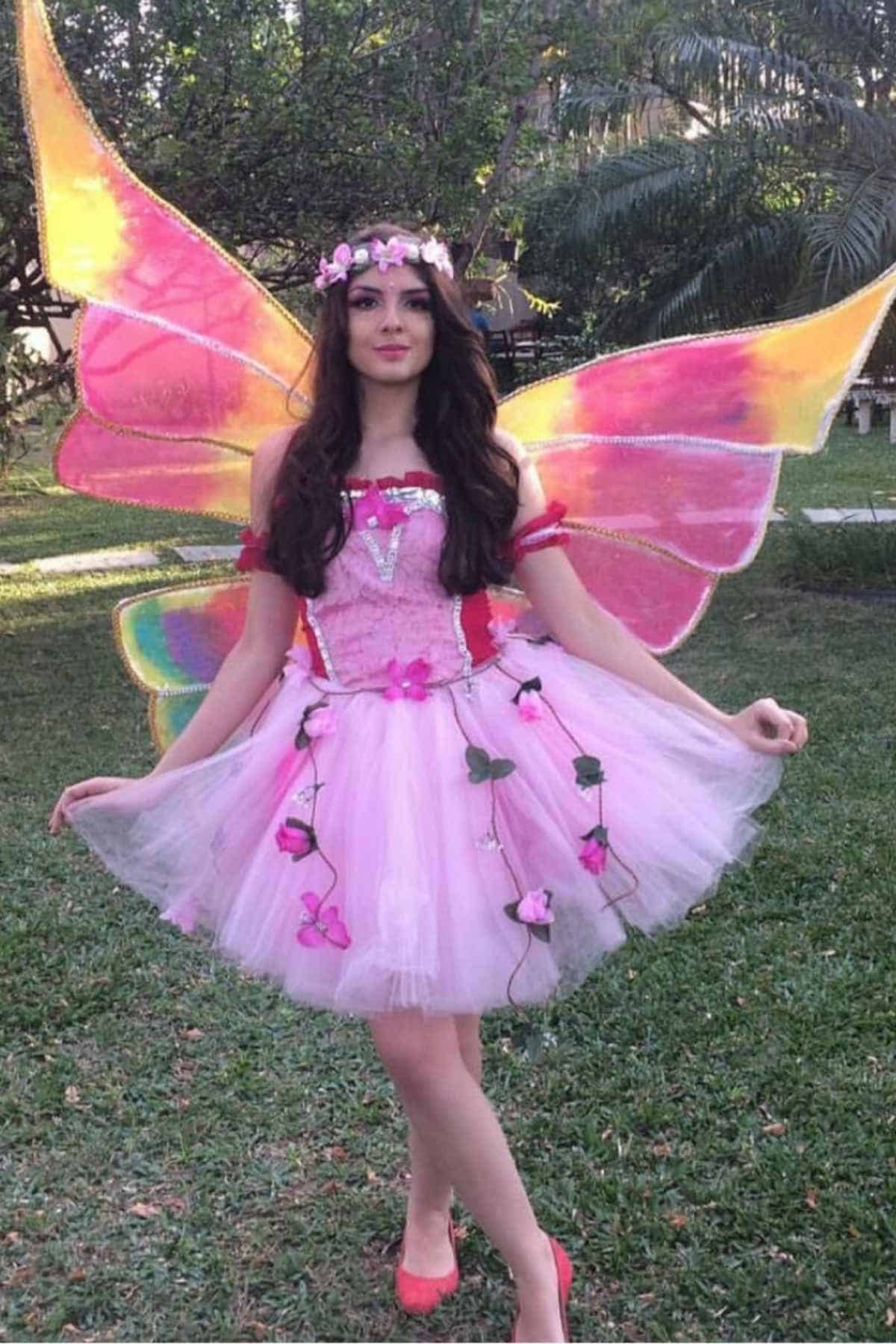A person in a pink fairy costume with large colorful wings and a floral crown stands on grass in a garden.