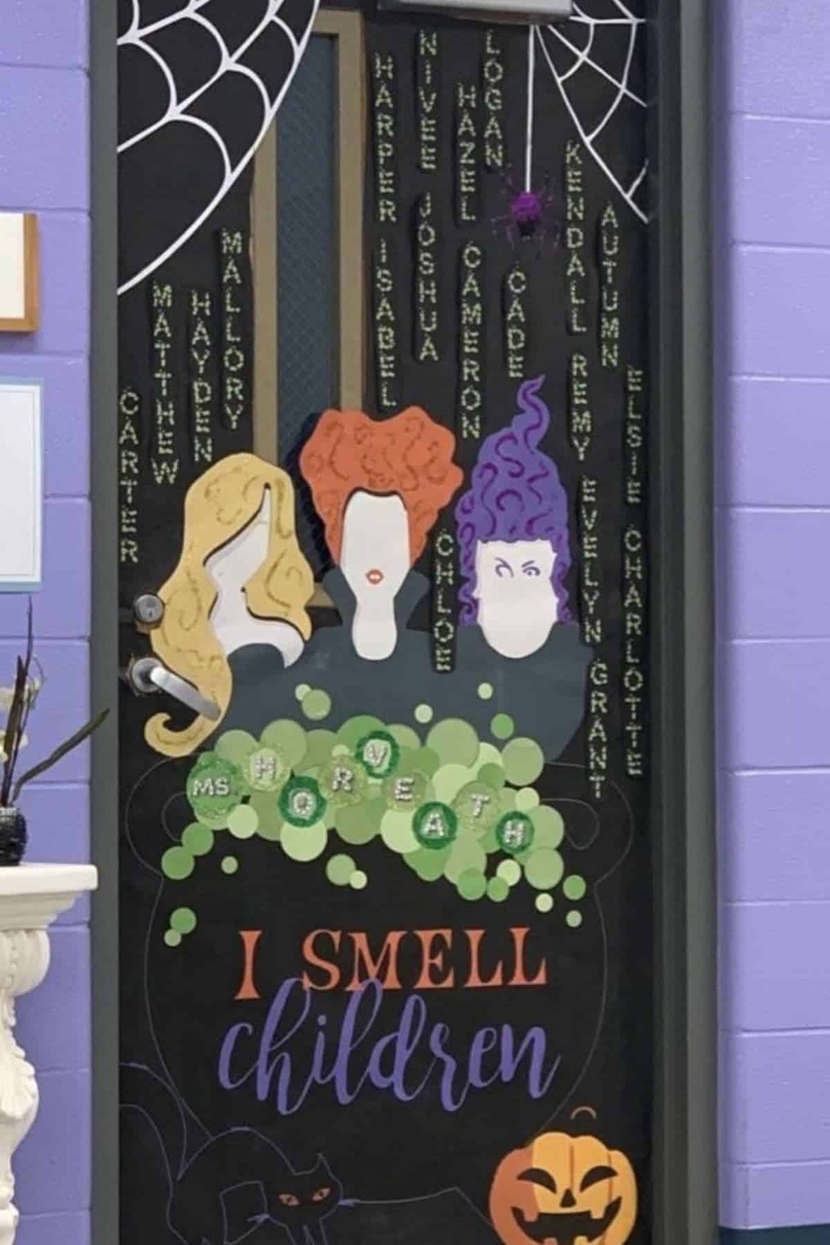 Classroom door decorated with three witch silhouettes, green bubbles, student names, spider webs, and a pumpkin. Text reads: “I SMELL children.”.