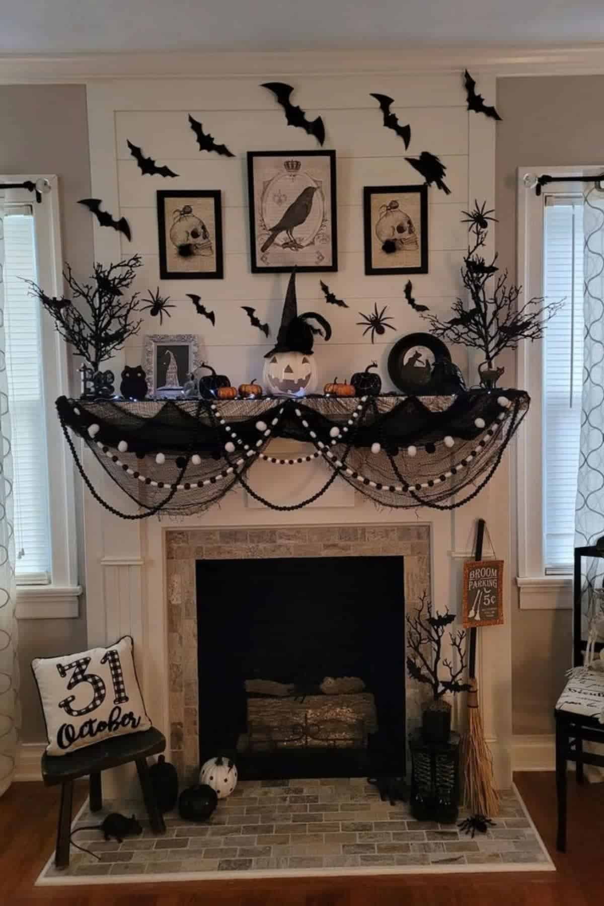 A fireplace mantel decorated for Halloween with black bats, a witch hat pumpkin, framed art, garlands, small pumpkins, fake trees, and a “31 October” sign.