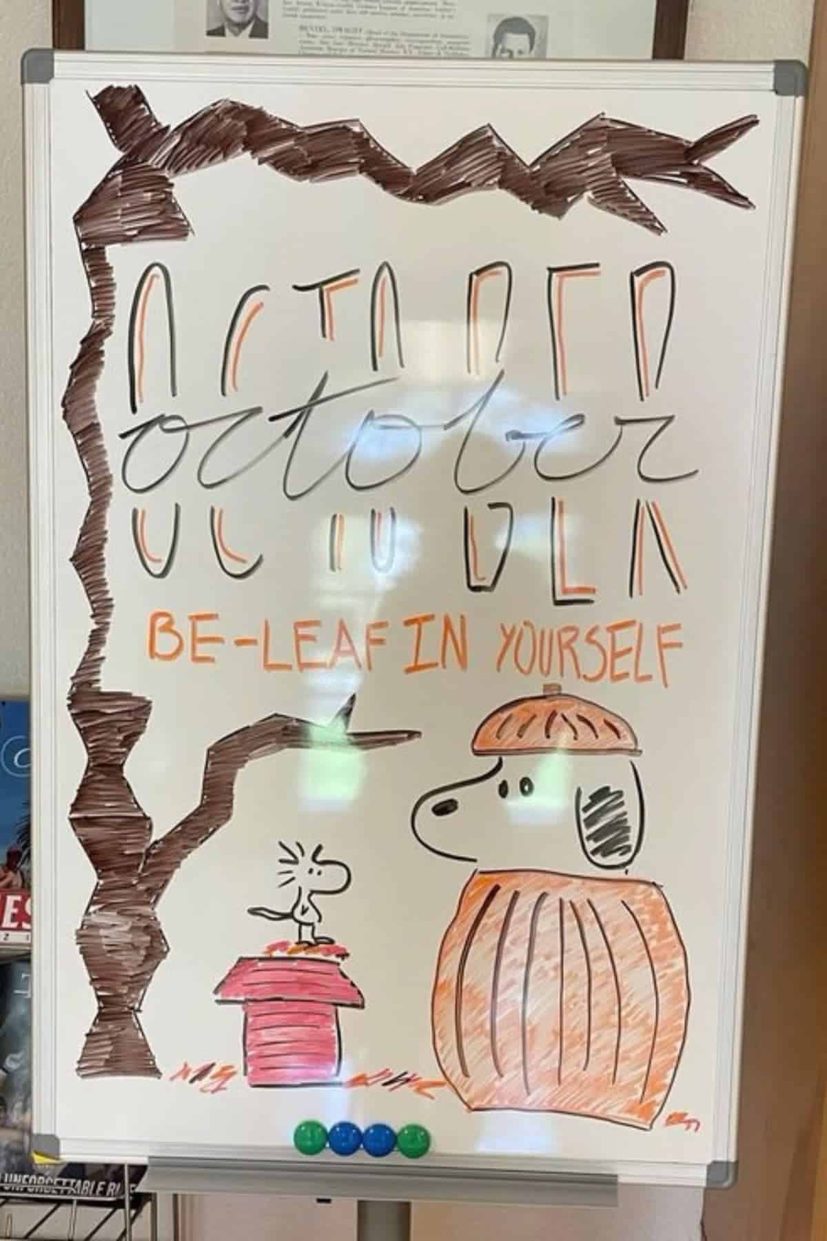 A whiteboard with “October” written in large letters, a dog and bird in autumn attire, trees, and the phrase “BE-LEAF IN YOURSELF” in orange marker.