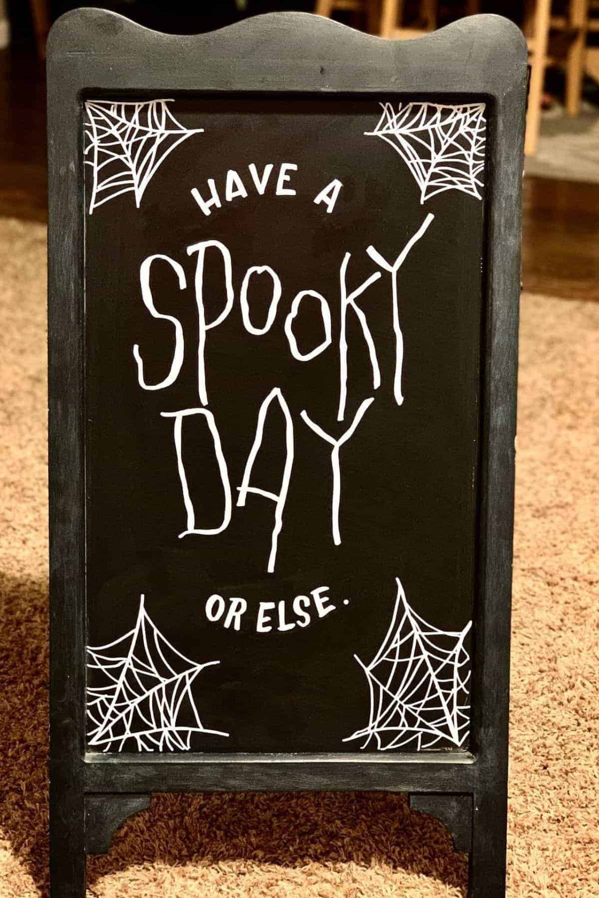 A black chalkboard sign reads “Have a Spooky Day or else.” with white spider web drawings in each corner.