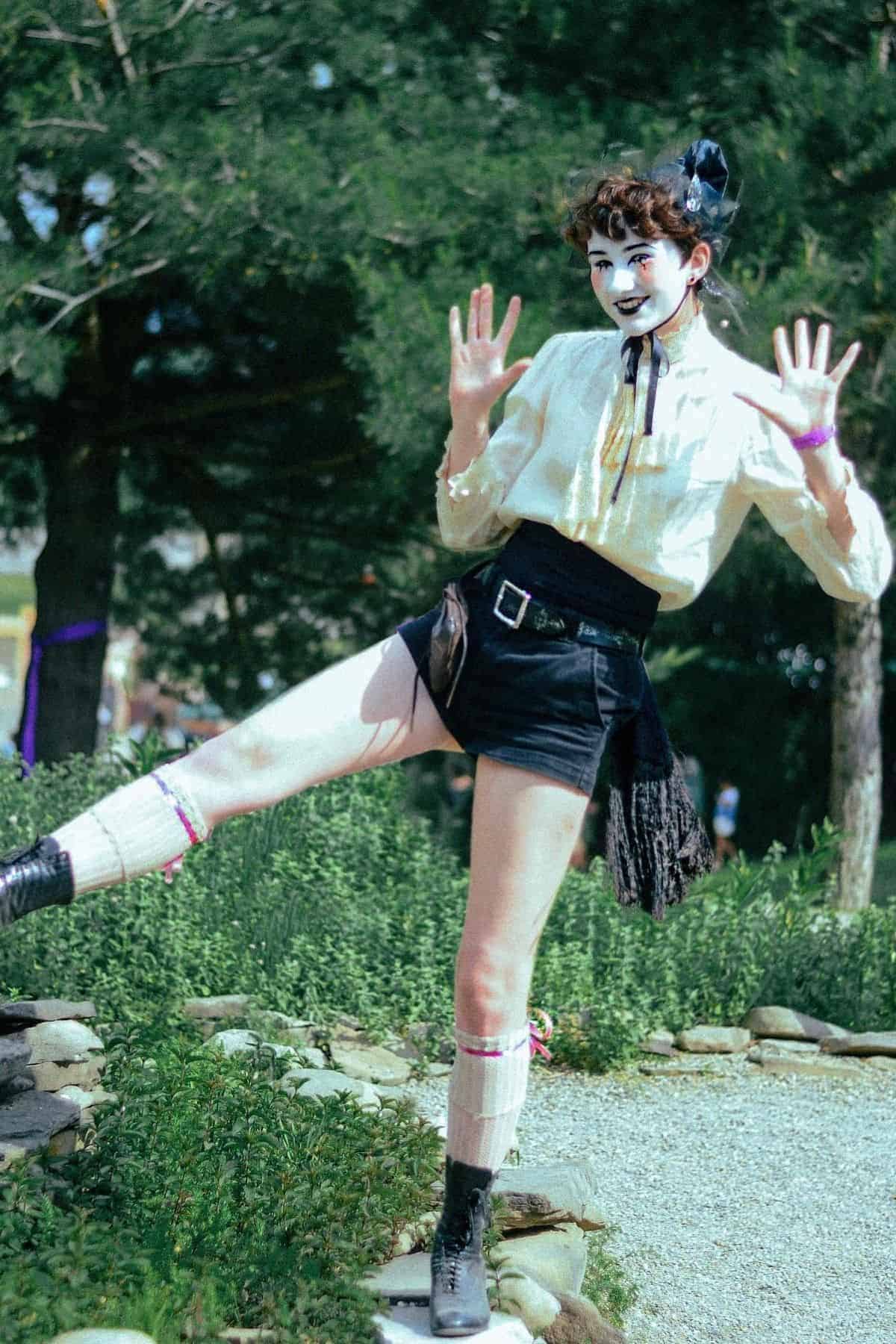 Person in theatrical makeup, white blouse, black shorts, and boots poses outdoors with one leg raised, hands up, and greenery in the background.