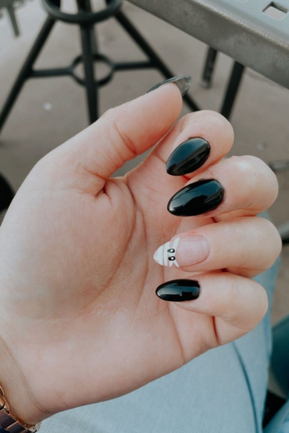 A hand with long, almond-shaped nails painted black, except for the ring finger, which has a mummy design with white wraps and two black eyes.