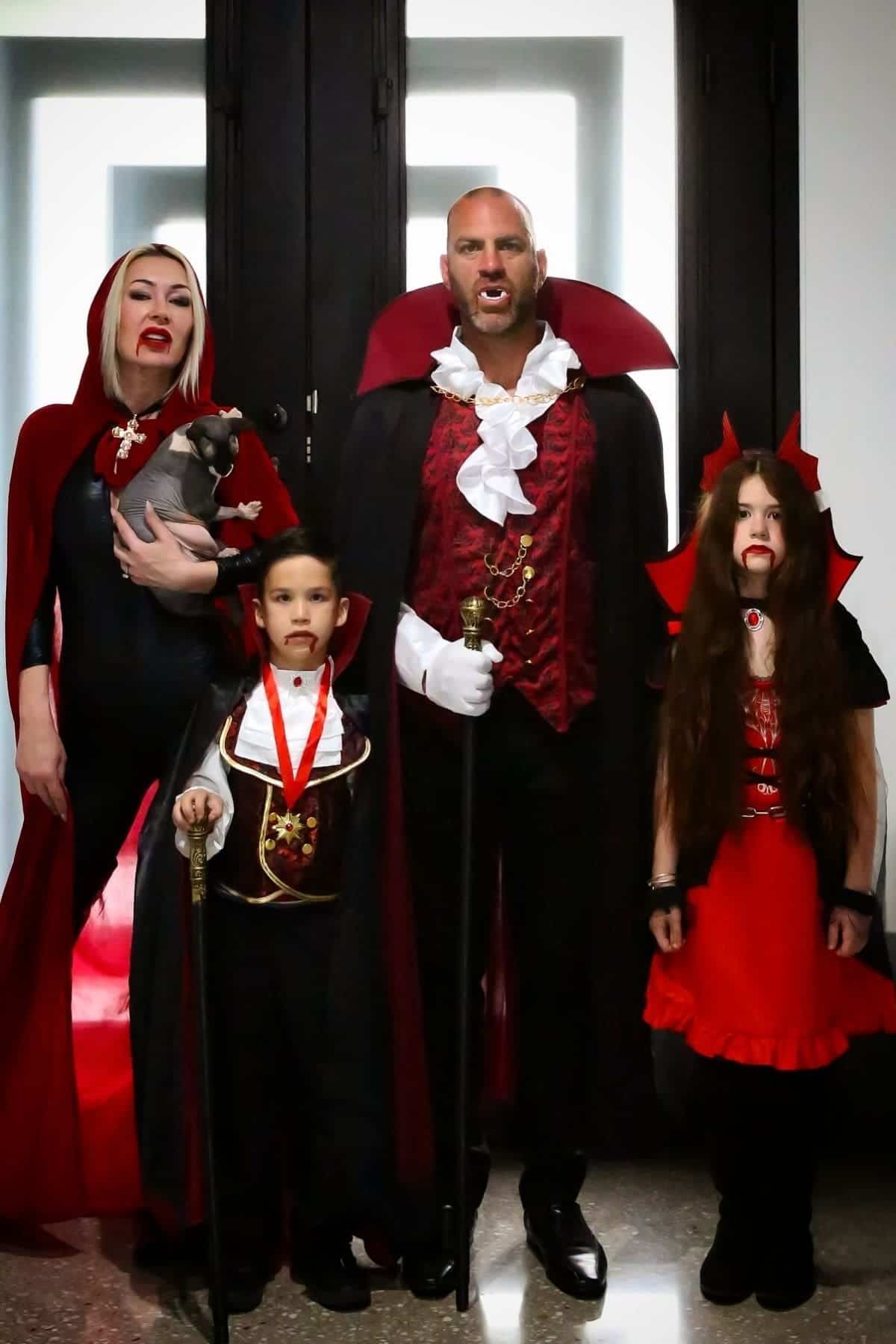 Four people and a dog dressed in matching vampire costumes stand indoors in front of dark double doors, all facing the camera.