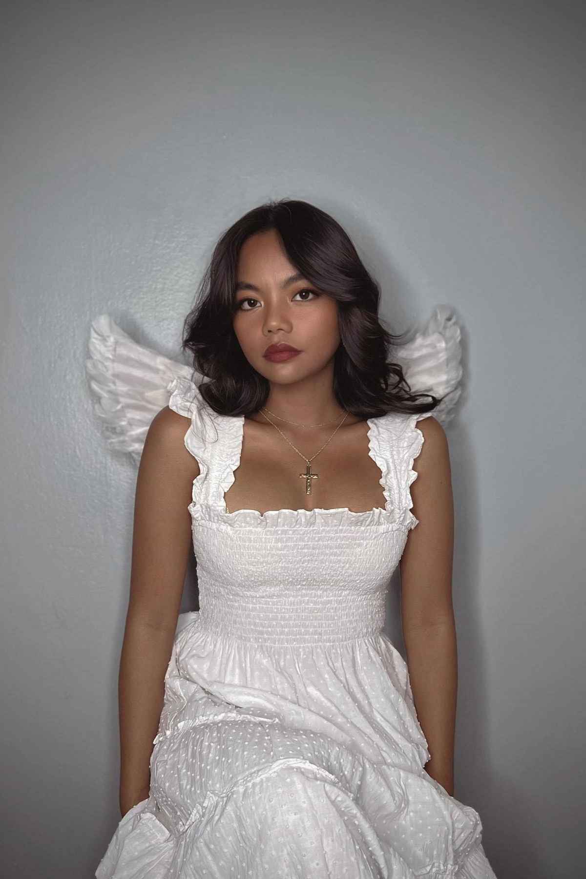 A woman in a white dress with a gold cross necklace poses against a gray wall, with white feathered angel wings visible behind her.