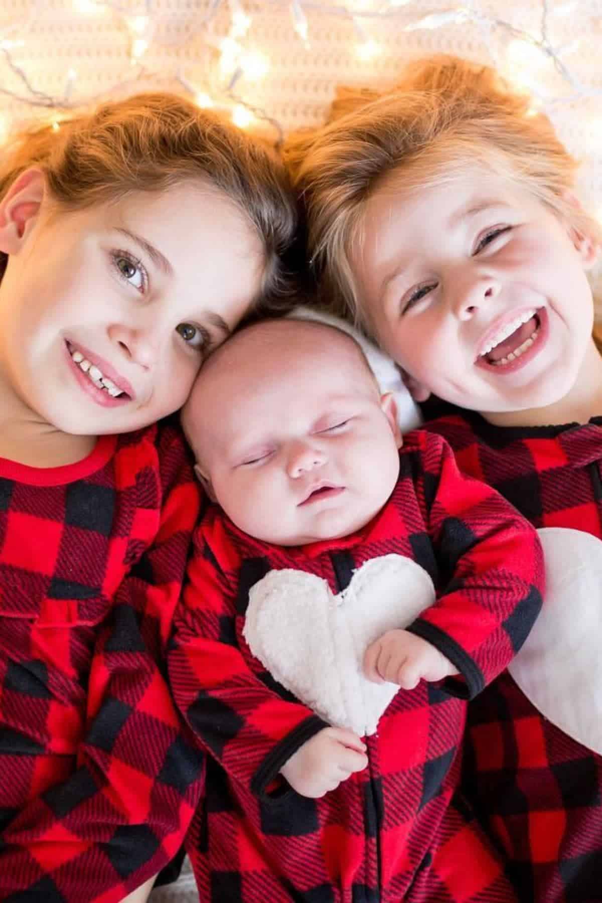 Three children in red and black plaid pajamas with white hearts lie side by side, smiling, with string lights in the background.